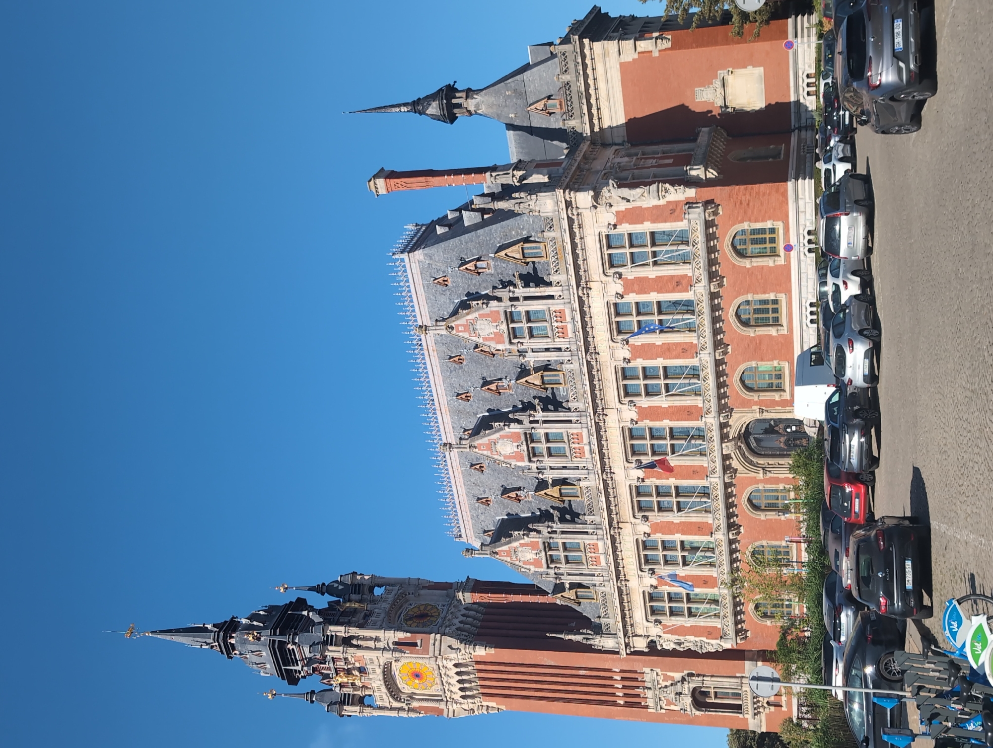 The image shows the exterior of the Calais City Hall, a grand brick building with a prominent clock tower, set against a clear blue sky. The architecture is ornate, featuring multiple gabled roofs, decorative stonework, and large windows. French flags are visible flying from the building. In the foreground, a parking lot is filled with various cars. Several bicycles, part of a rental system, are parked on the left side of the image. The building's details suggest it is the Hôtel de Ville of Calais, France, given its distinctive architectural style and the presence of French flags. The scene appears to be a typical daytime view of a public building in a European city, with everyday elements like parked cars and a bike share system.