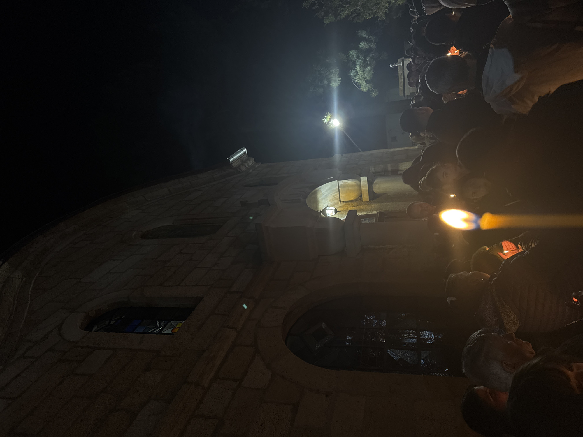 At night, a crowd of people holding candles stands in front of a stone building, which is lit by a street lamp. The building's architecture features windows and stone blocks. The scene appears to be taking place in Byala Slatina, Bulgaria, given the context.