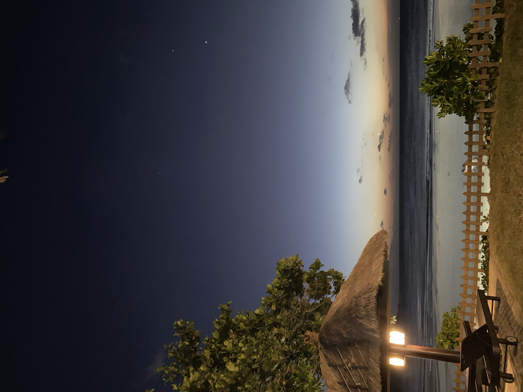 The image depicts a serene coastal scene at twilight or very early morning in Kuta Selatan, Indonesia. The original image is rotated 90 degrees counter-clockwise; when viewed correctly, the sky occupies the upper portion, transitioning from a deep indigo with visible stars at the top to a lighter blue, orange, and pink hue along the horizon line. A few dark clouds are scattered across the lighter part of the sky.
Below the horizon, the dark, calm ocean stretches across the middle ground, gently meeting a narrow strip of sandy beach. On the right side of the image, a light-colored wooden picket fence separates a grassy area from the beach. Green foliage, likely bushes or small trees, dot the landscape, with one prominent, leafy tree extending into the frame from the bottom left.
In the bottom right foreground, a structure with a thatched roof, resembling a large beach umbrella or a small hut, is visible. Attached to its support pole are two warm, glowing artificial lights, casting a yellow light on the immediate area. Part of a dark outdoor chair or piece of furniture is also visible near the lights and fence.
No people or visible text are present in the image. The scene suggests a peaceful, relaxing atmosphere at a beachside location, possibly set up for evening dining or leisure by the sea.