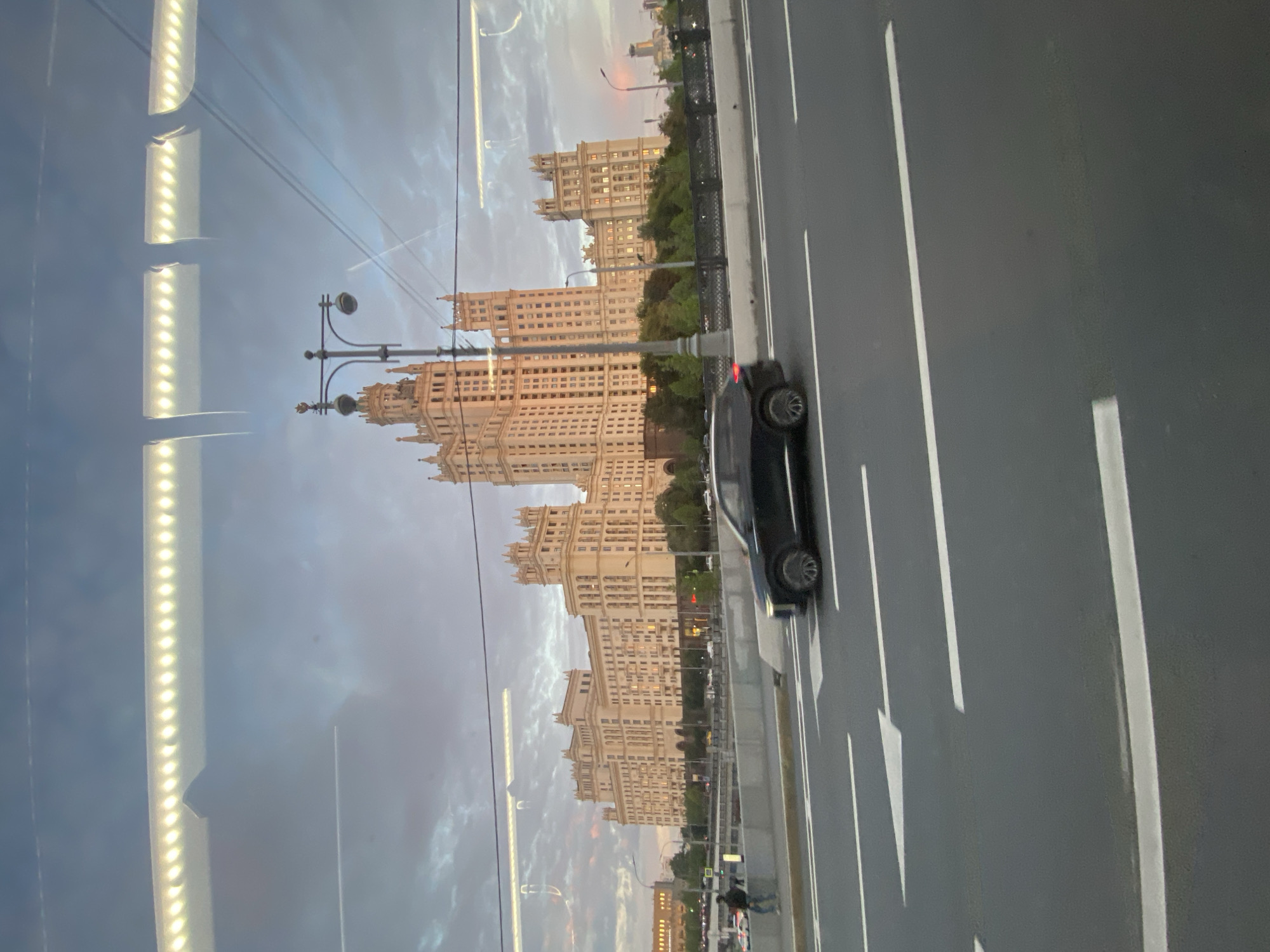 The image depicts a view of the Stalinist skyscraper known as the Kudrinskaya Square Building in Moscow, Russia, taken from a moving vehicle. The building, a prominent landmark, is a large, ornate structure with multiple towers and intricate architectural details, illuminated by artificial lights. In the foreground, a black SUV is driving on a multi-lane road. The road has clear white lane markings, including directional arrows. Beyond the road, a concrete barrier separates it from a pedestrian walkway, where a few individuals are visible, one walking. Trees and greenery are situated behind the barrier, leading up to the base of the skyscraper. The sky is overcast with a mix of grey and pinkish clouds, suggesting it is either sunrise or sunset. Reflections of lights, possibly from the vehicle's interior, are visible on the surface from which the photo was taken, suggesting it was shot through a window.