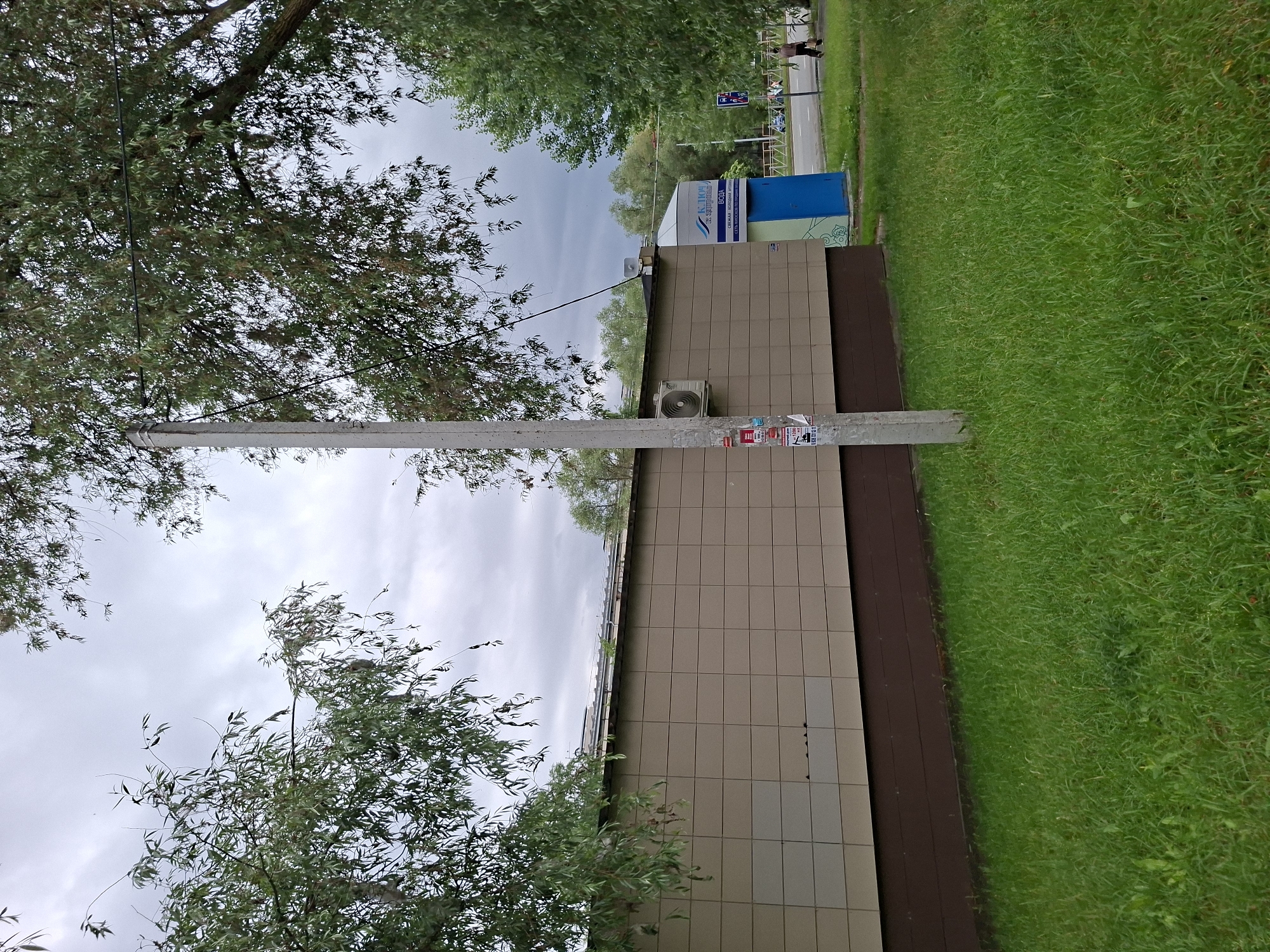 A cloudy, overcast day is depicted in an outdoor scene. In the foreground, a well-maintained lawn of green grass stretches across the frame. A concrete utility pole stands prominently on the left side of the image, adorned with several pasted notices. Behind the pole and to its right, a light brown, tiled building extends horizontally. An air conditioning unit is mounted on the side of this building.
To the right of the building, a blue kiosk with white text on its upper section is visible. The text is in Russian, and appears to say "КЛЮЧ К ЗДОРОВЬЮ" and "ВОДА" indicating a water dispenser or similar service. Further to the right, a paved road runs parallel to the building. A person is walking on the sidewalk next to the road, and a traffic sign with a pedestrian symbol is visible on the right. Lush green trees with dense foliage frame the upper portions of the scene, their branches reaching across the sky, partially obscuring the clouds. The overall impression is of a functional, possibly commercial or public area within a city.