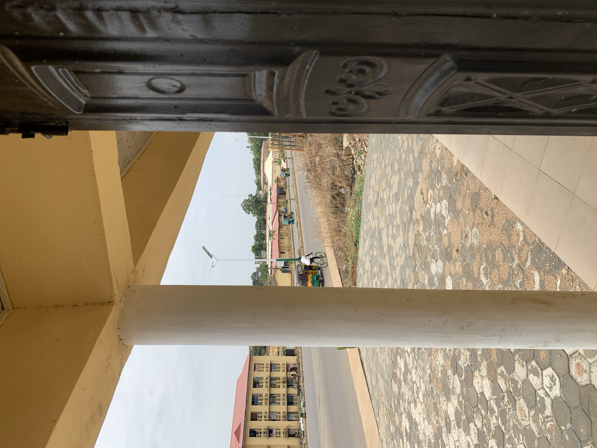 The image shows a street scene in Jos, Nigeria, viewed through an open doorway. A concrete column partially obstructs the view. A paved area with a hexagonal pattern is visible to the right of the column. Across the street, there is a building with a red roof and multiple windows. A person is riding a bicycle alongside a motorized tricycle. The sky is overcast. The doorway is dark and ornate.