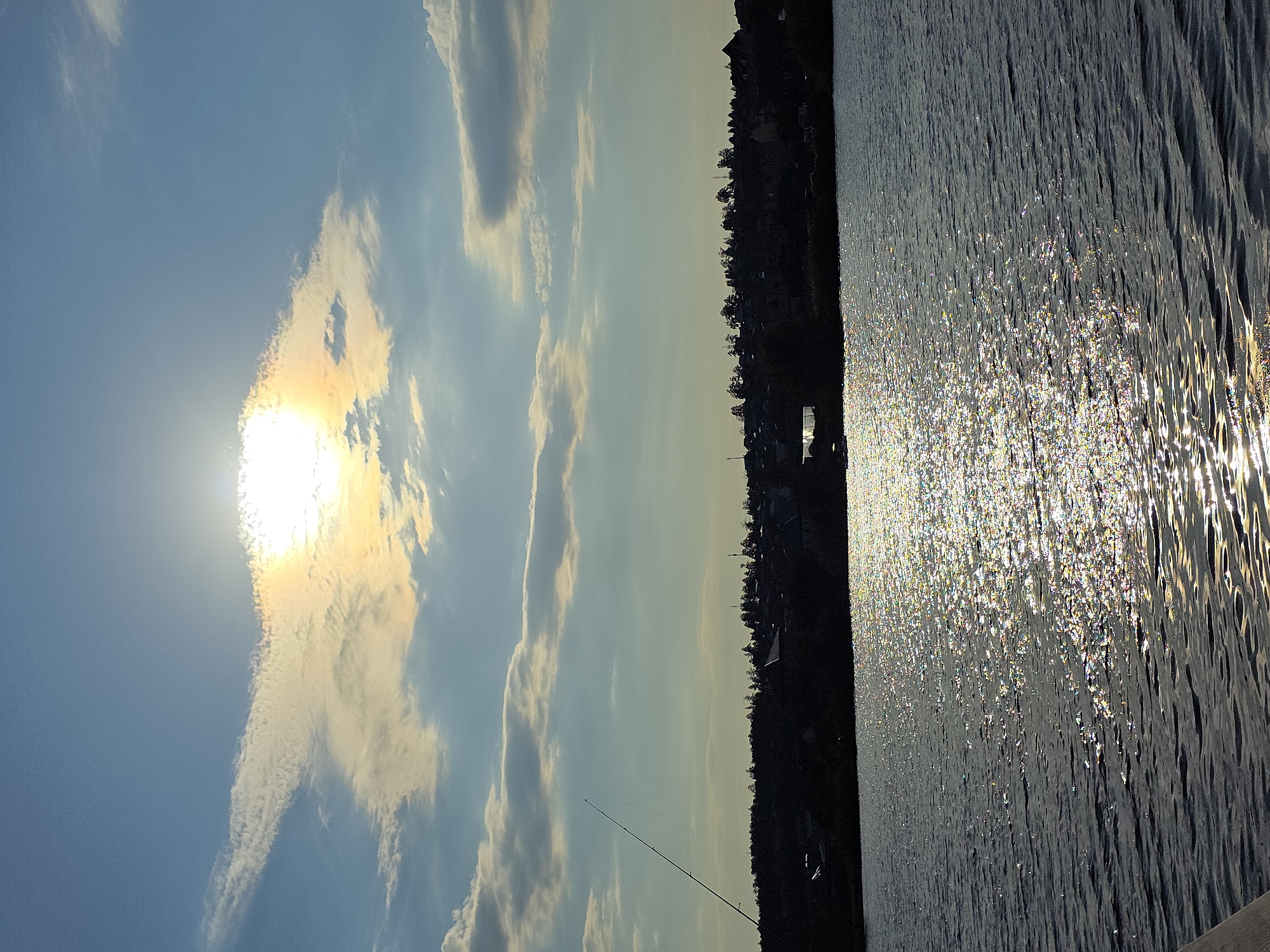 The image depicts a serene lake with a silhouetted treeline and buildings along the horizon under a bright, partly cloudy sky. The sun, partially obscured by clouds, casts a shimmering reflection on the water's surface. A fishing rod extends into the frame from the left, suggesting a recreational activity. The scene is set against a backdrop of a clear blue sky with scattered white and golden-tinged clouds. The overall impression is one of tranquility and natural beauty, likely captured during the late afternoon or early evening. The location is identified as Lake Turgoyak in Miass, Russia.