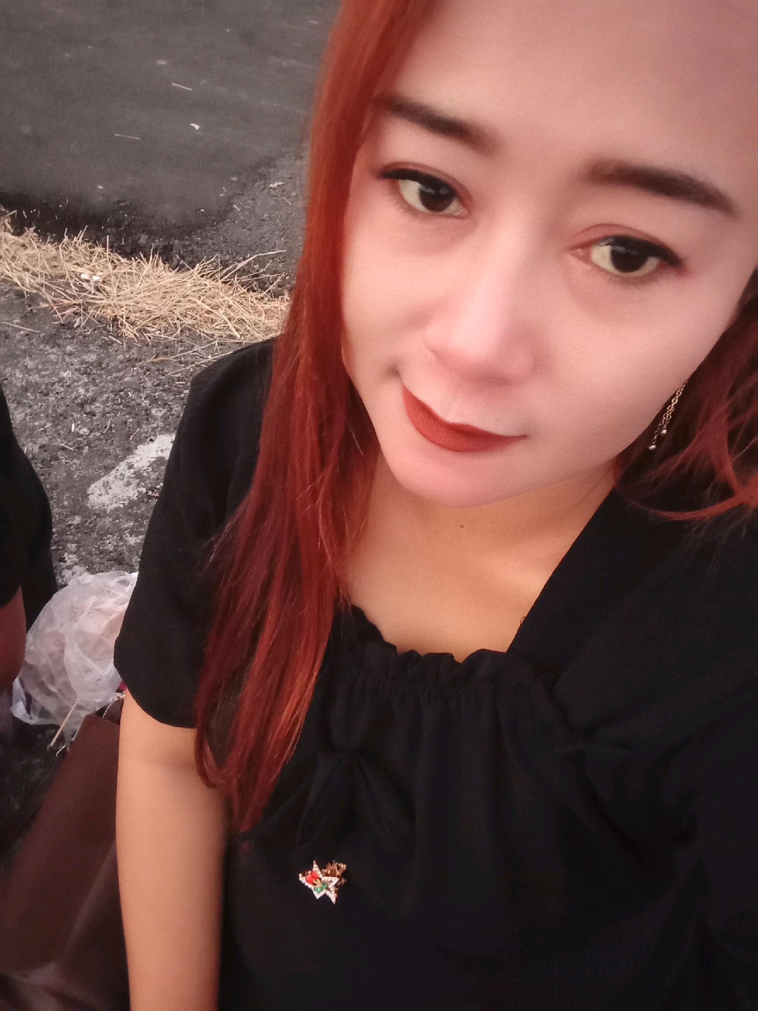 In a close-up, selfie-style photograph, a woman with auburn hair and red lipstick smiles at the camera. She wears a black top adorned with a star-shaped brooch. To her left, a pile of dried grass sits on a dark, paved surface, possibly a road or pathway. A white plastic bag is visible in the lower left corner. The background is out of focus, with a general appearance of an outdoor, possibly rural, setting. The lighting suggests an overcast day. No specific landmarks or buildings of Desa Ngargorejo, Indonesia are visible.