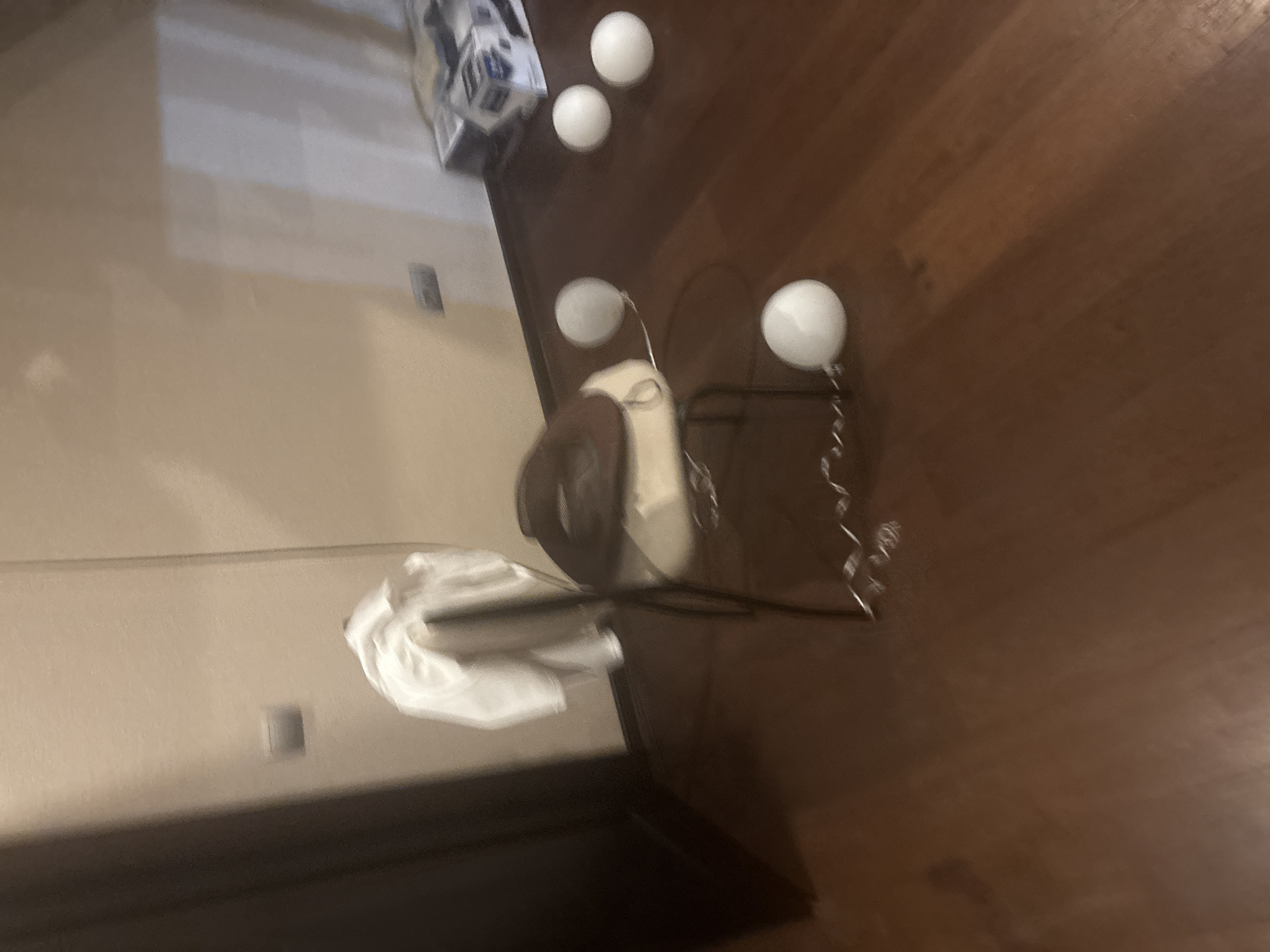 The image shows a chair with a white garment draped over its back, and a hat on its seat. There are several white spherical objects on the wooden floor in front of the chair, some of which appear to be balloons. In the background, a wall with light-colored paint is visible, along with what looks like a power outlet. To the right, there are boxes stacked on the floor. The lighting is dim, suggesting an indoor setting. The location is specified as Orekhovka, Russia.