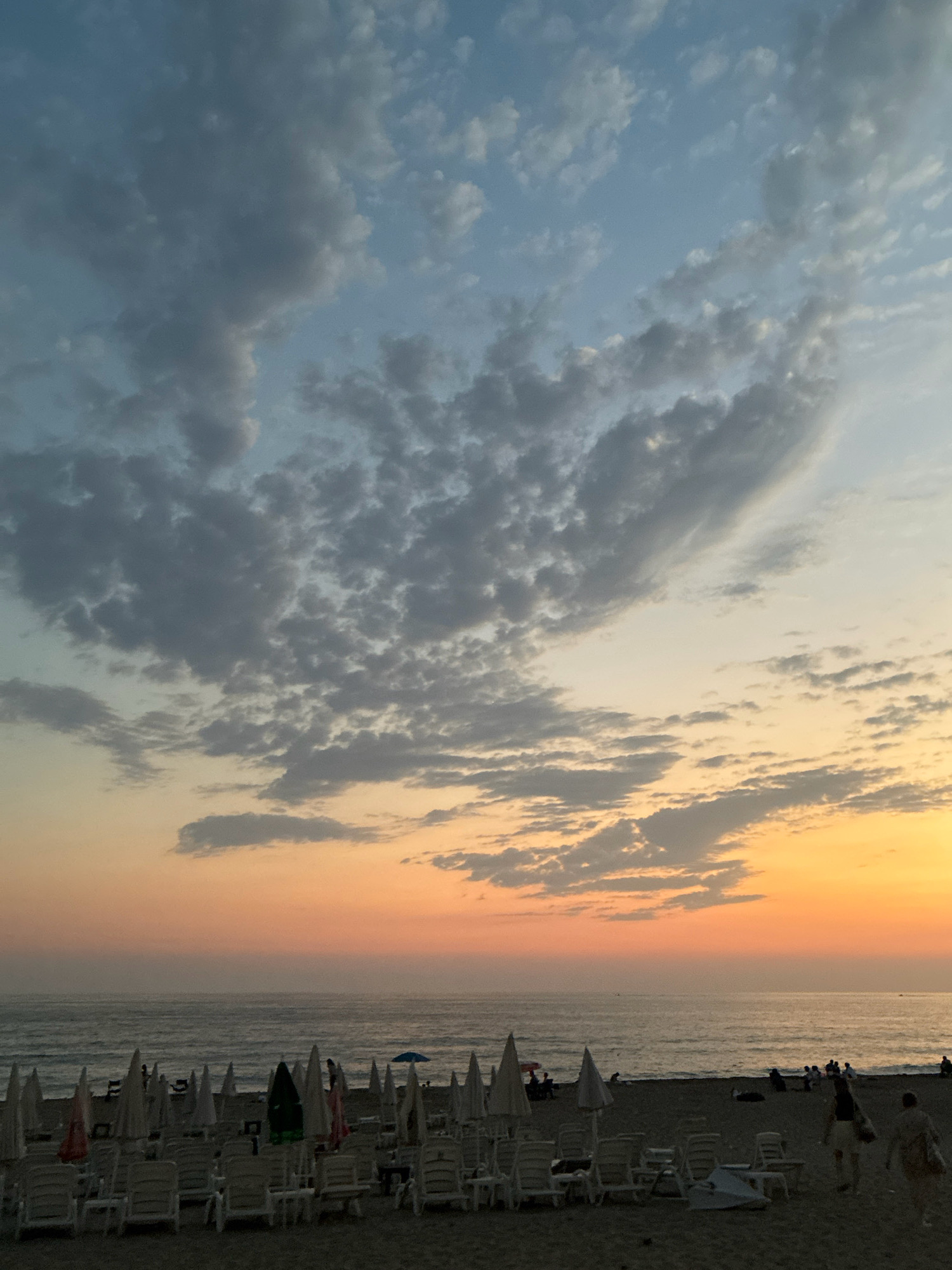 The image shows a beach in Alanya, Turkey, at sunset. The sky is filled with a mix of blue and orange hues, with clouds scattered across the horizon. The sea is calm, with gentle waves lapping at the shore. On the beach, numerous empty lounge chairs and closed umbrellas are arranged in neat rows, indicating the end of the day. Several people are visible, some walking along the shoreline, others sitting or standing near the water. A few individuals are further back on the sand, near the loungers. The overall atmosphere is peaceful and serene, capturing the end of a day at the beach.