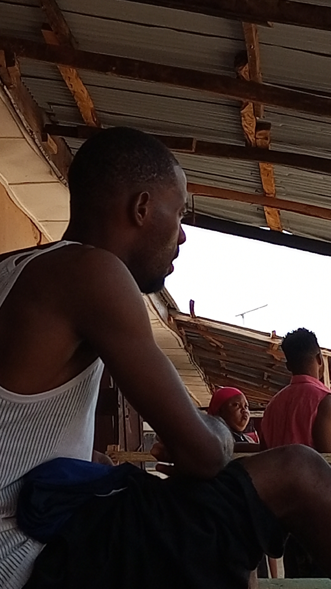 The image captures a partially obscured outdoor scene in Sabon Tasha, Nigeria. An African man wearing a white and grey striped tank top and dark shorts sits in the foreground, obscuring the view of other people. Behind him, two other people are visible. A woman wearing a pink head covering looks in one direction, and a man wearing a pink shirt faces the opposite direction. The setting appears to be a covered, outdoor area with a corrugated metal roof supported by wooden beams, suggesting a public space or a commercial area. A television antenna is also visible.