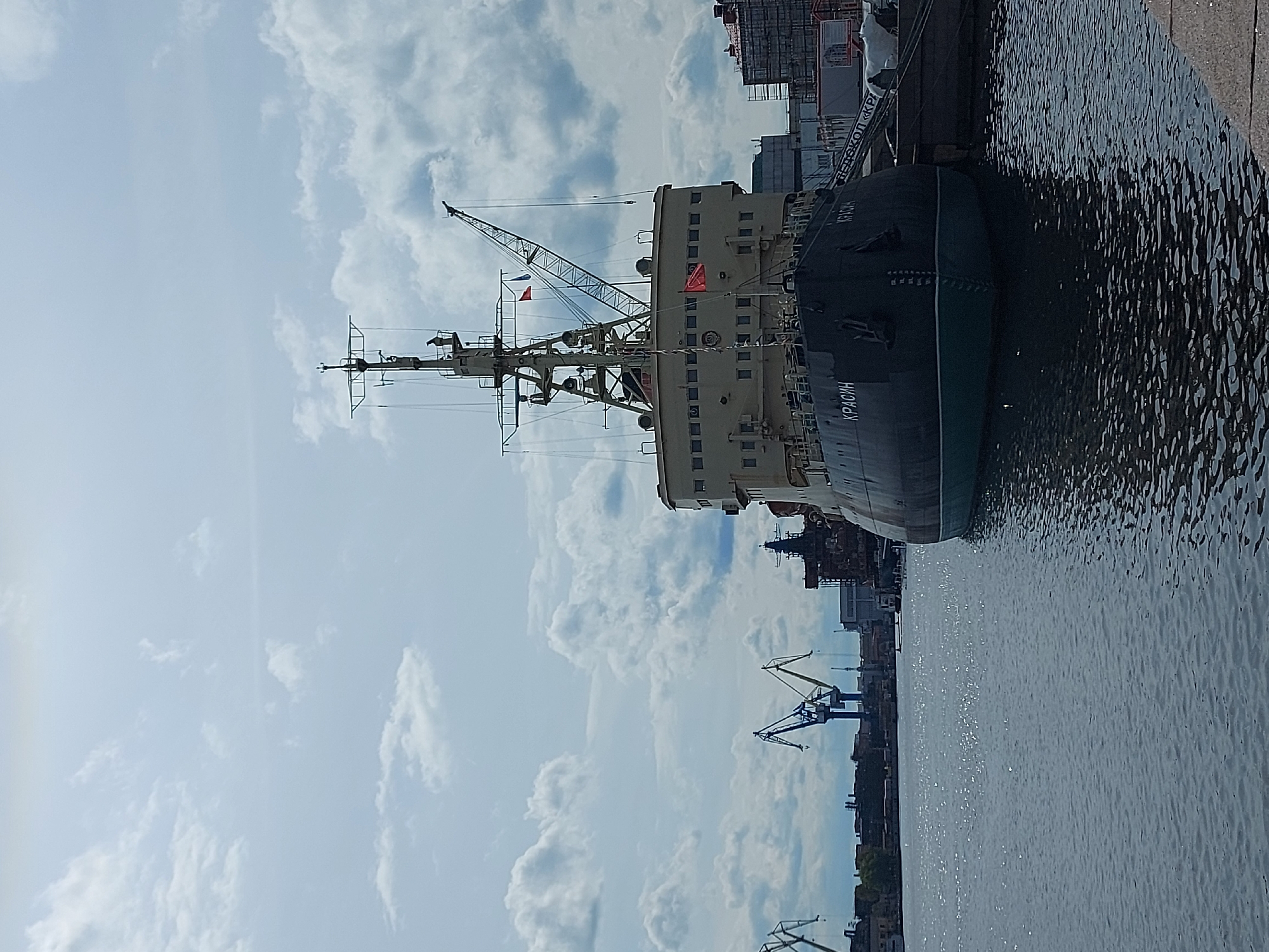 The image shows the icebreaker Krasin docked in Saint Petersburg, Russia. The ship is positioned along a quay, with the water of the Neva River visible on the right. The ship's bow is facing towards the viewer, and the name "КРАСИН" (KRASIN) is visible on its side. The superstructure of the ship, including a crane and masts, rises against a cloudy sky. In the background, other buildings and cranes are visible, indicating an industrial or port area. The image is taken during daylight, and the lighting suggests a partly sunny day.