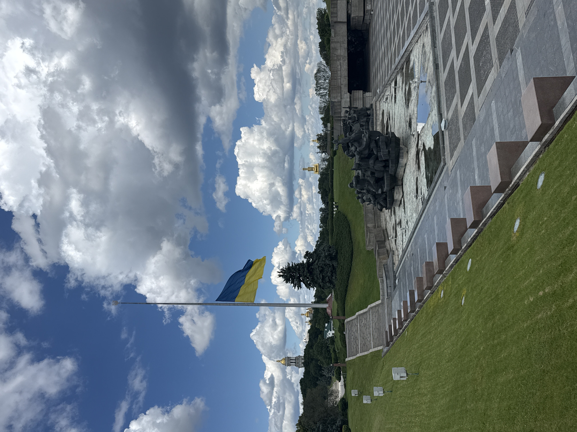 This image depicts a wide-angle, eye-level outdoor view of the Motherland Monument and the National Museum of the History of Ukraine in the Second World War complex in Kyiv, Ukraine. The scene is dominated by a clear blue sky with scattered, fluffy white clouds. In the foreground and mid-ground, a large, manicured green lawn slopes down towards a paved area with a large monument featuring abstract human figures. A wide set of stone stairs leads up the grassy slope.
A prominent, tall flagpole stands centrally in the image, flying the blue and yellow flag of Ukraine. The flag is partially unfurled, creating a dynamic visual element against the sky.
In the background, several golden-domed religious buildings, likely churches or cathedrals, are visible on a tree-covered hill. The Motherland Monument itself, a colossal stainless steel statue, is partially obscured by the flag and the surrounding greenery, but its iconic silhouette can be discerned. To the right of the monument complex, a modern bridge or overpass is visible.
The overall atmosphere is one of solemnity and national pride, enhanced by the presence of the national flag and the monumental statues. The environment is well-maintained, with lush grass and clean paved areas, suggesting it is a public space designed for commemoration and reflection. No people are visible in the image. The lighting suggests it is daytime, likely late morning or early afternoon, with the sun casting soft shadows.