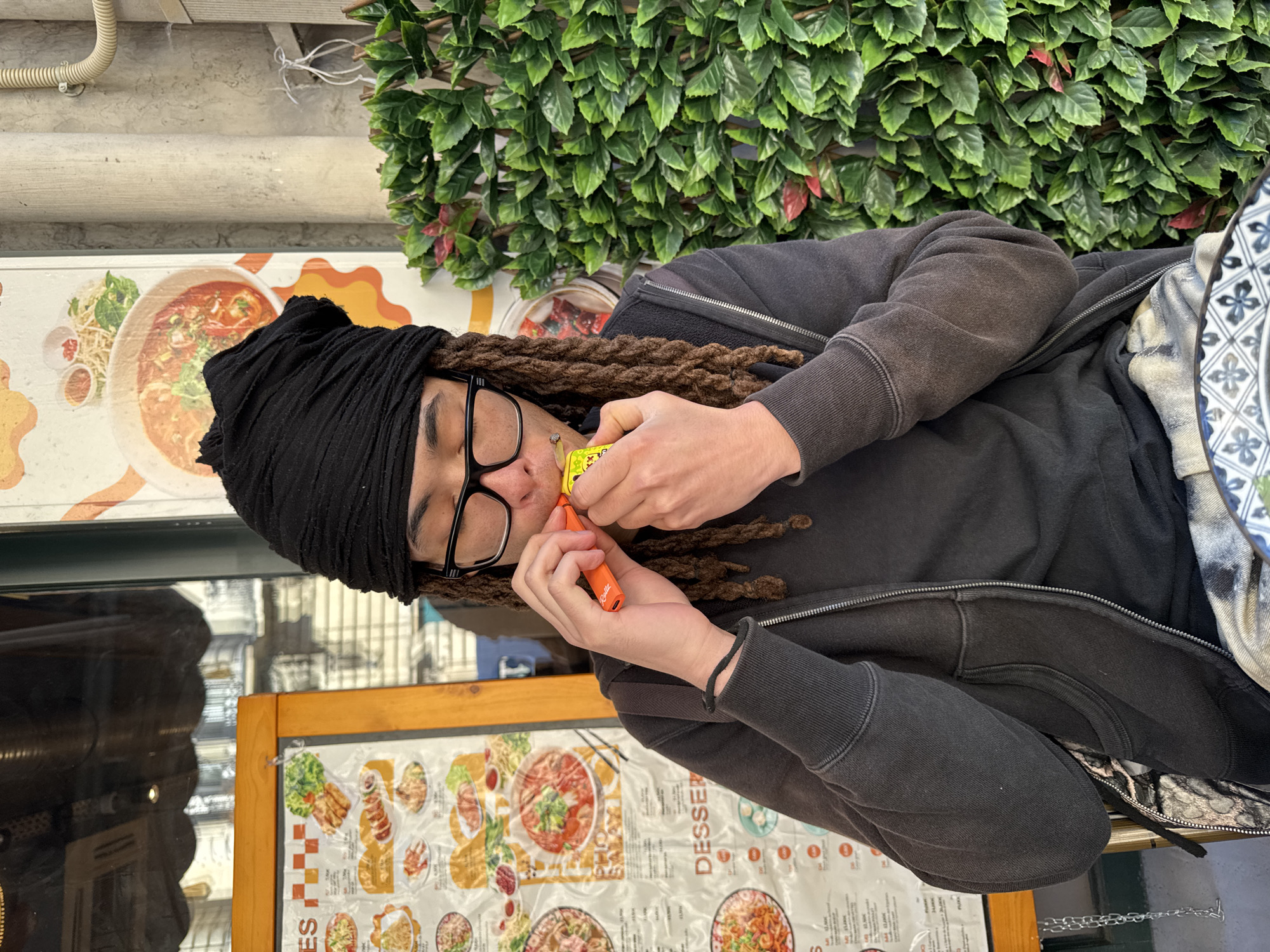 This image, likely taken in Paris, France, captures a young person seated outdoors at what appears to be an Asian restaurant during the daytime. The subject, seen from the chest up, has dark dreadlocks, wears a black head covering resembling a turban, and black-framed glasses. They are dressed in a dark grey or black hooded zip-up sweatshirt.
The person is actively engaged with two small items held in their hands, close to their face. Their right hand holds an orange cylindrical device, possibly a vape pen, on which the brand name "Riedler" is visible. In their left hand, they hold a small yellow package adorned with cartoon-like characters, from which a small, hand-rolled item, suggestive of a cigarette or joint, is protruding. Their expression is focused and neutral.
The background features a vibrant wall covered in dense artificial green foliage with some reddish accents, creating a decorative outdoor setting. Above this, a concrete wall with white piping can be seen. To the person's right, a patterned blue and white fabric is visible. Several menu boards are present in the scene; one above the person displays various dishes typical of Vietnamese cuisine, including pho. Another wooden-framed menu board below the person clearly shows the word "DESSERT" among other illegible text. Reflections in a nearby glass surface reveal glimpses of surrounding buildings. The natural light indicates it is daytime.