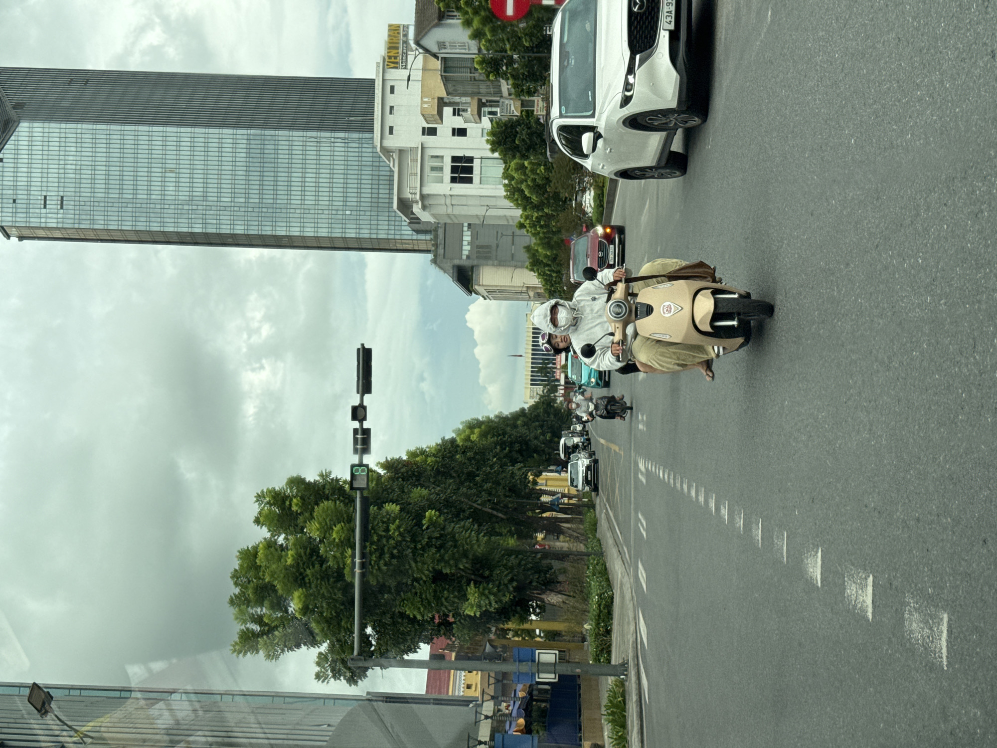 The image depicts a street scene in Thuận Hóa Ward, Vietnam. In the foreground, a beige electric scooter carries two individuals. The driver, wearing a white hooded jacket and a face mask, is in the center, with a passenger behind. The scooter is moving on a paved road with dashed white lane markings. To the right of the scooter, a white Mazda CX-5 SUV is visible. Further down the road, a red car and other vehicles are present, along with a line of trees and buildings. A tall, modern glass skyscraper dominates the background, with other urban structures and a cloudy sky. A traffic light, showing a green signal, is mounted on a pole to the left. The overall environment suggests a busy urban street with ongoing traffic.