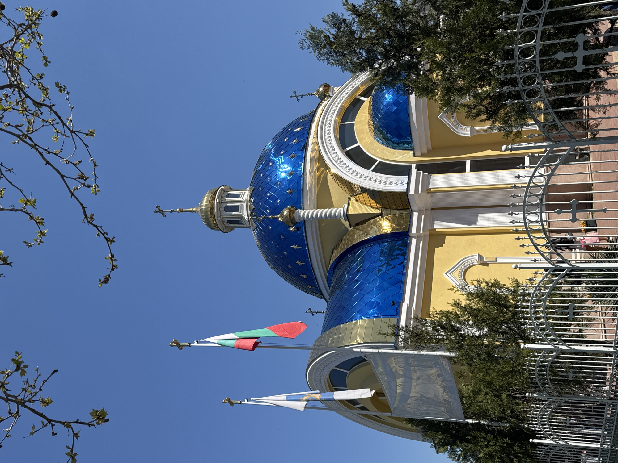 The image shows the Orthodox Church "All Saints" in Plovdiv, Bulgaria, viewed from a low angle. The church features a blue, star-studded dome with gold accents. Bulgarian flags are displayed in front of the church, alongside trees and a metal fence. The sky is clear and blue.