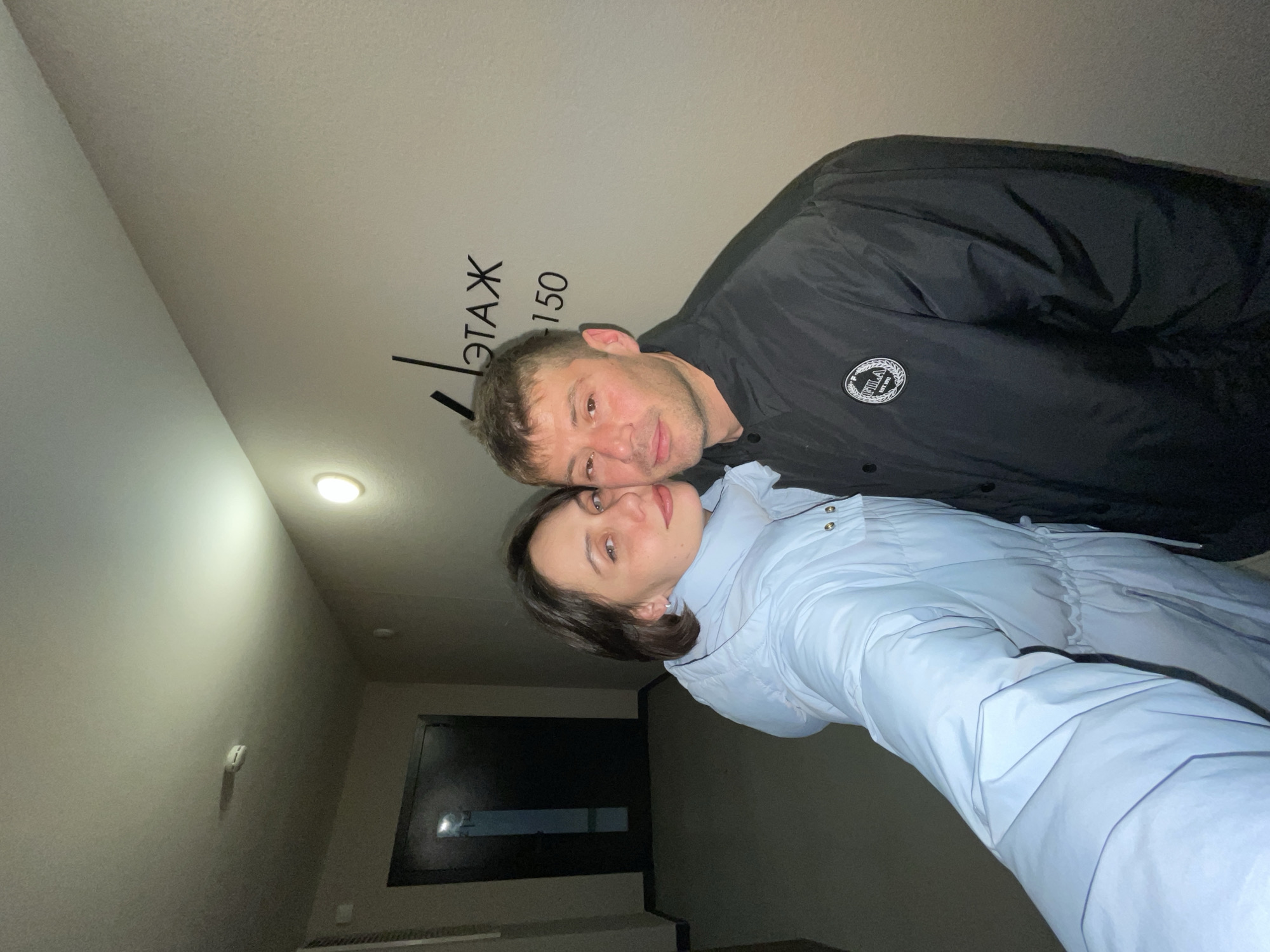 Two individuals, a man and a woman, are posing closely together in what appears to be an indoor setting. The man is wearing a black jacket with a round logo, and the woman is wearing a light blue puffer jacket. They are positioned in front of a wall with the words "ЭТАЖ 150" written in black, indicating they are likely on the 150th floor of a building. A doorway is visible in the background. Based on the information from the writing "ЭТАЖ", the location is in the city of Pervomaysky, Russia.