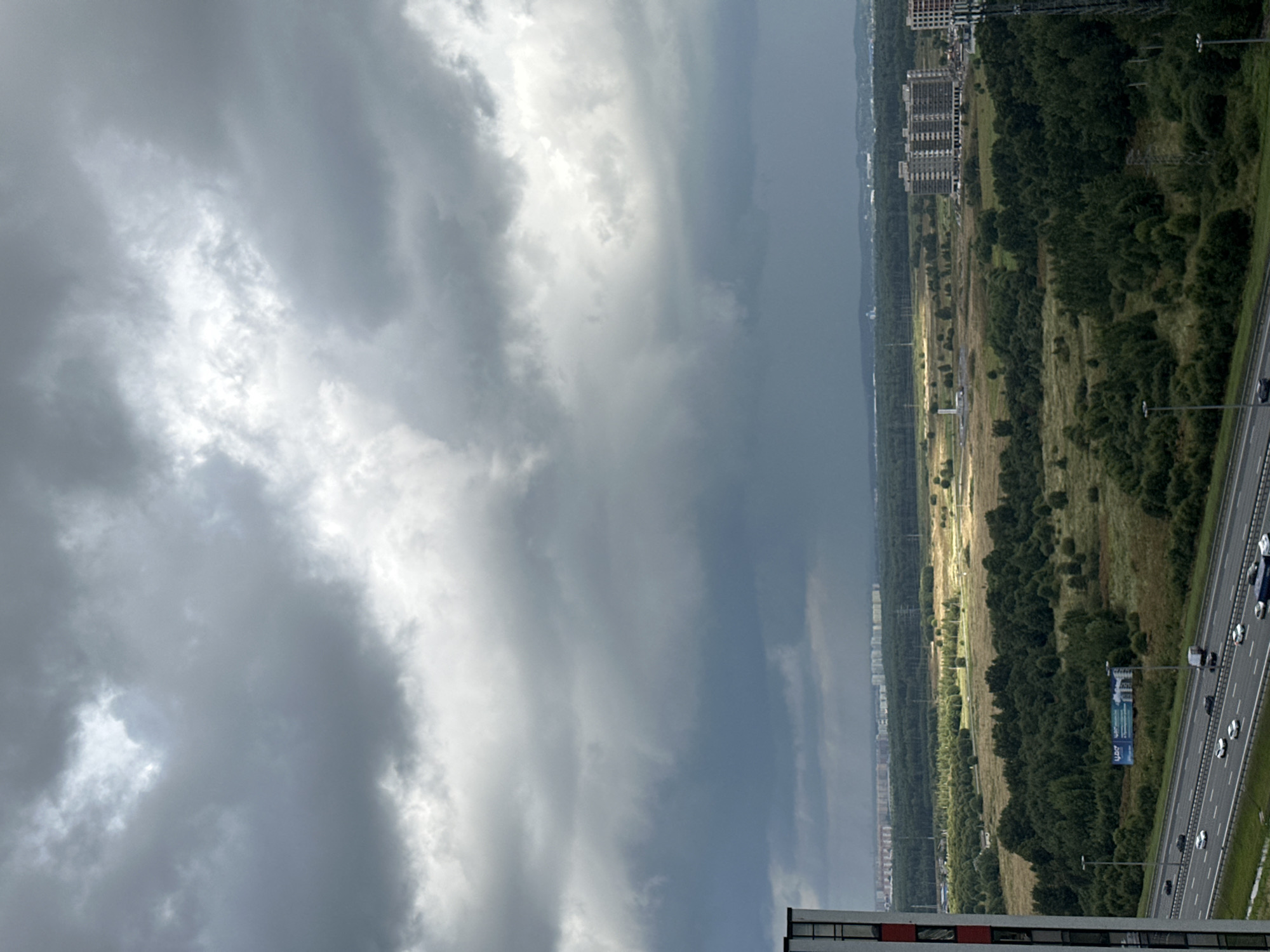 The image captures an overcast sky over a suburban landscape in Otradnoye, Russia. In the foreground, a multi-lane highway with moderate traffic runs horizontally across the frame. Cars, including several white sedans and a dark-colored SUV, are visible on the road. To the left, the edge of a tall building with a red and white facade is partially visible.
The middle ground features a vast expanse of green fields and scattered clusters of trees. A large billboard is positioned along the side of the highway. Further into the distance, more dense forests are visible, interspersed with patches of drier vegetation.
On the right side of the image, a large, multi-story apartment building with a grid-like pattern of windows is prominent. Beyond this building, a more distant cityscape with numerous buildings can be seen, stretching towards the horizon. The sky is dominated by heavy, dark gray clouds, suggesting impending or ongoing precipitation, with a faint patch of lighter sky visible towards the center. The overall scene conveys a sense of a somewhat gloomy day in a developing urban area surrounded by natural landscapes.
