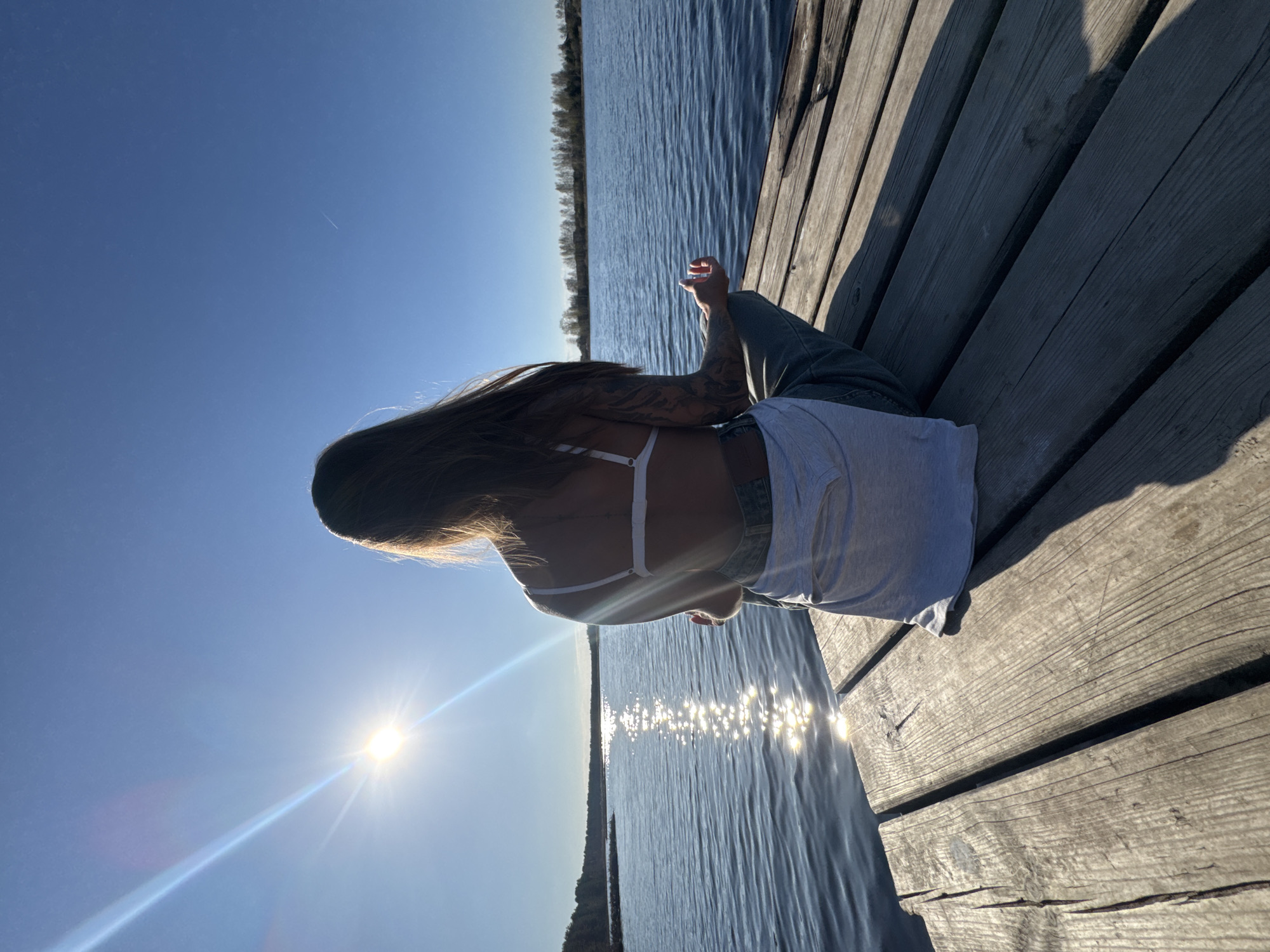The image depicts a woman sitting on a wooden dock, facing away from the camera. She is wearing a white tank top, blue jeans, and has a tattoo on her left arm. Her long hair is blowing in the breeze. The background consists of a calm lake and a clear blue sky. The sun is shining brightly, reflecting off the water. The location is near Bol'shoye Kozino, Russia, based on likely landscape characteristics.