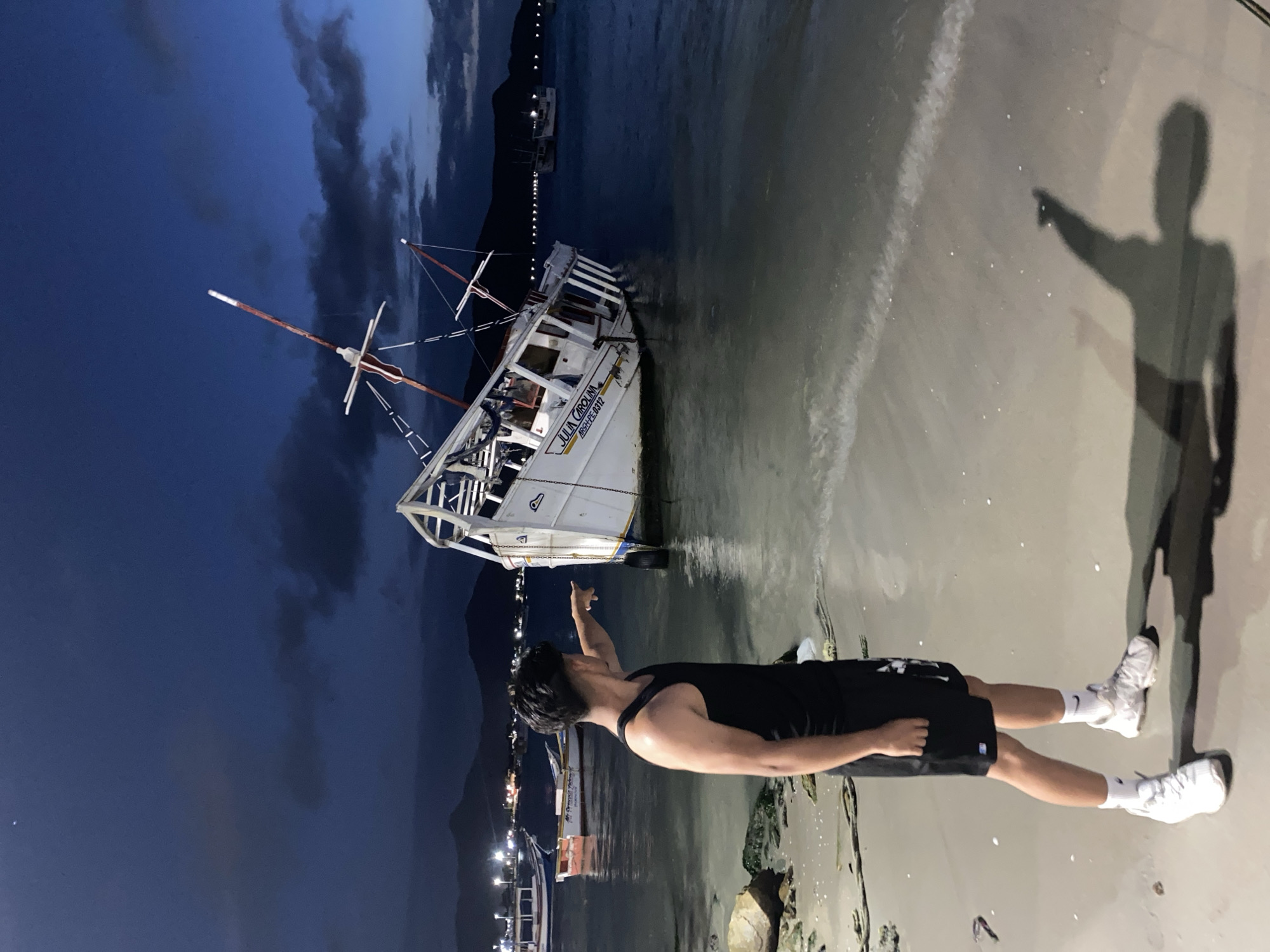 A man in black shorts and a black tank top stands on a sandy beach, pointing towards a white ship beached on the shoreline in Juan Griego, Venezuela. The ship, named "Julia Carolina" is listing slightly to the side. The scene is at night, with lights visible along the coastline in the background. The sky is dark blue with some clouds. A shadow of the man is cast on the sand next to him.