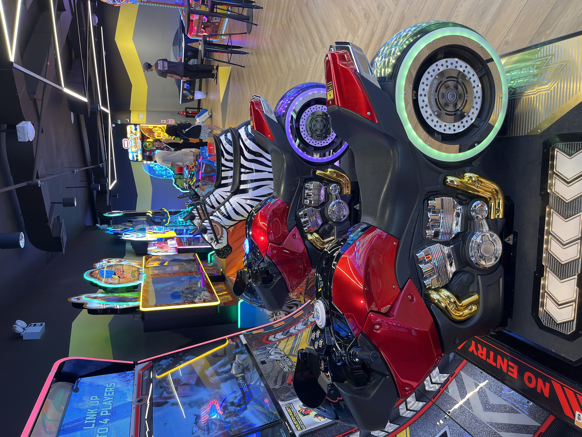 This media file captures an interior scene of an arcade, likely in Kuta, Indonesia, showcasing a row of "Speed Rider" motorcycle arcade games. The games are designed with vibrant colors and intricate details, featuring sleek red and black motorcycle bodies, chrome accents, and illuminated wheels with green and purple lighting. Some of the games have unique zebra-patterned seats.
In the background, other arcade games with colorful displays and neon lighting are visible, indicating a lively and entertaining environment. Several people are present, some standing and observing the games, while others appear to be playing or waiting. One person is wearing a grey t-shirt with some text that is partially visible, and another has a white t-shirt. The lighting in the arcade is a mix of bright overhead fluorescent lights and the colorful glow from the game machines, creating a dynamic atmosphere.
The visible text includes "SPEED RIDER," "DEUS ex MACHINA," "INDUSTRY FIRST! Next level Drifting Expe," "NO ENTRY," and "LINK UP TO 4 PLAYERS." These texts suggest that the game is a racing or drifting simulator that can be played by multiple people, and it is a notable innovation in the industry. The overall scene conveys a sense of fun and excitement typical of an arcade. There are no clear indicators of the time of day or weather conditions, but the indoor setting implies it could be any time.
