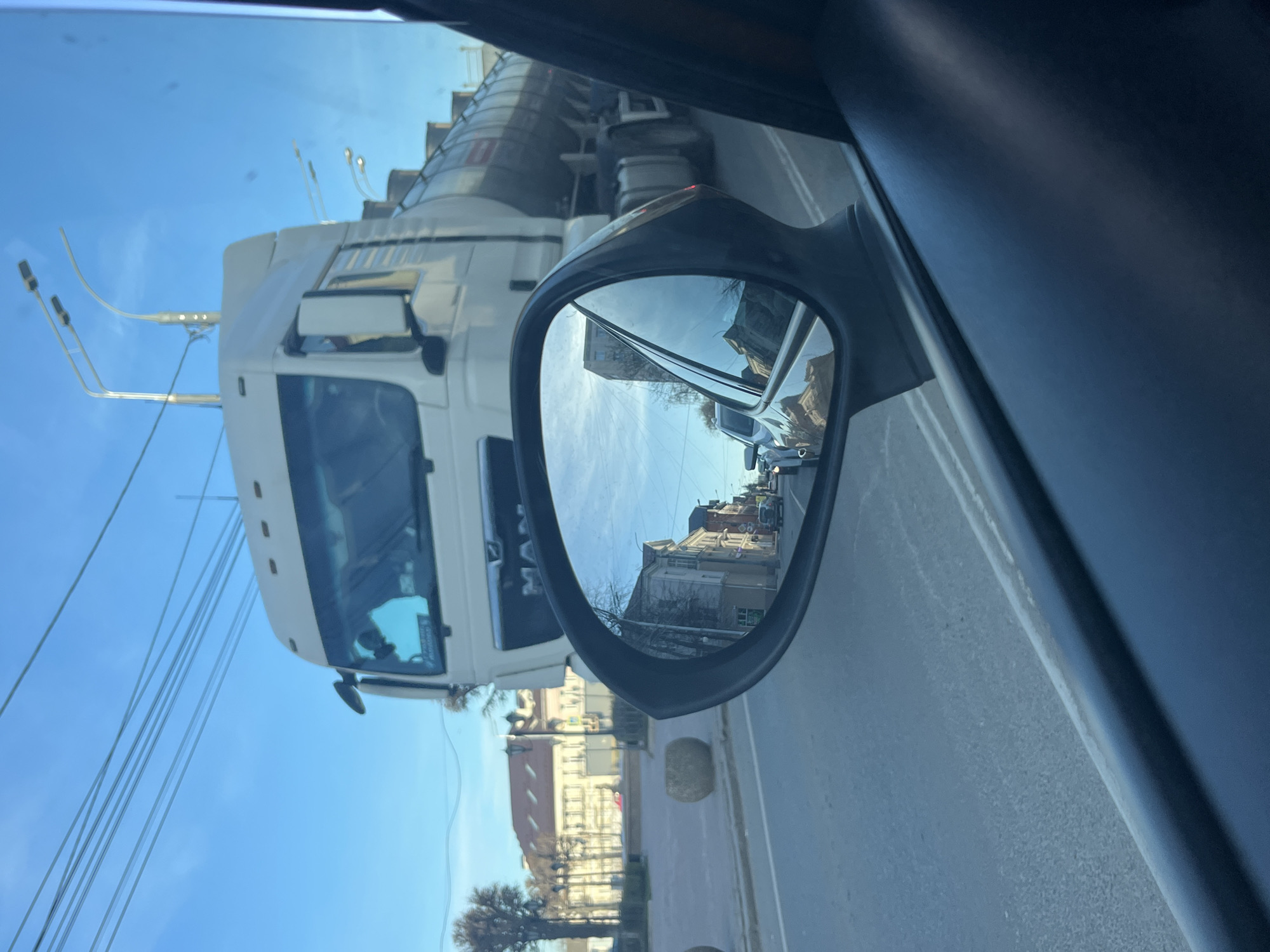 This media file captures a street view from inside a vehicle, likely a car, looking out the side window. The scene is oriented from the perspective of a front-seat occupant.
**Subjects:** A significant portion of the left side of the frame is occupied by a large white truck, possibly a semi-truck or tanker, positioned alongside the car. The car's side mirror is visible on the right, reflecting a detailed street scene. Through the car window and reflected in the mirror, multi-story buildings with classic architecture line the street, accompanied by bare trees. Overhead, numerous power lines or trolley lines crisscross a clear blue sky.
**Activity:** The car appears to be stationary or moving slowly in traffic, positioned next to the large truck on a city street.
**Notable details:** The weather is clear and sunny, indicated by the bright blue sky and the quality of light. The bare trees suggest a season like late autumn, winter, or early spring. A round, light-colored object, possibly a stone bollard, is visible on the roadside in the background. The overall impression is an urban environment in Klin, Russia.
**Visible text:** The word "MAN" is clearly visible on the grille of the large white truck.
