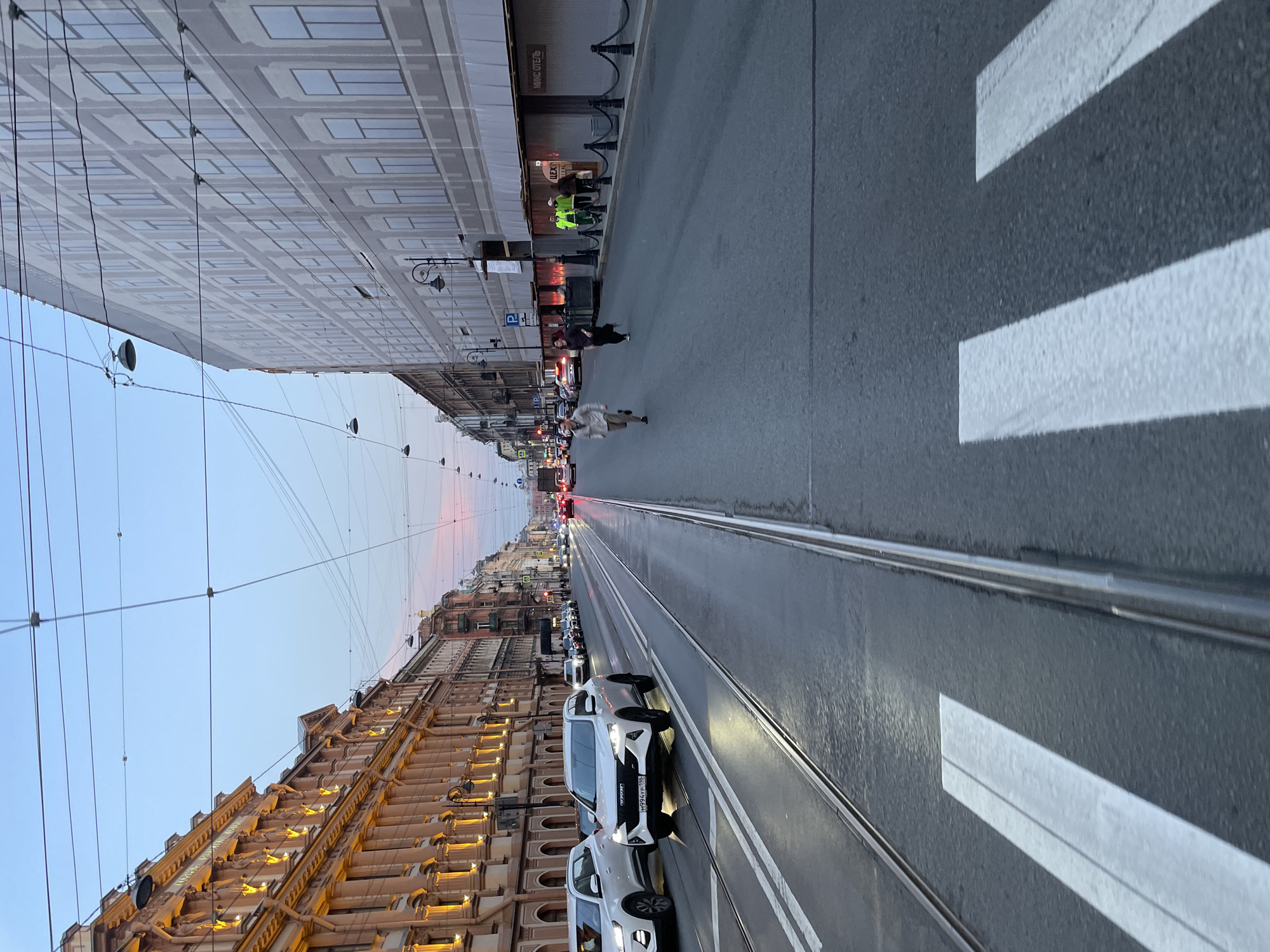 The image captures a street scene in Saint Petersburg, Russia. The road is divided by a tram track running down the center. White zebra stripes indicate a pedestrian crossing in the foreground. Several white cars are parked on the right side of the street. People are walking along the sidewalks and crossing the street. Buildings line both sides of the street, one with detailed architectural elements illuminated by lights. Overhead power lines are visible against the sky.