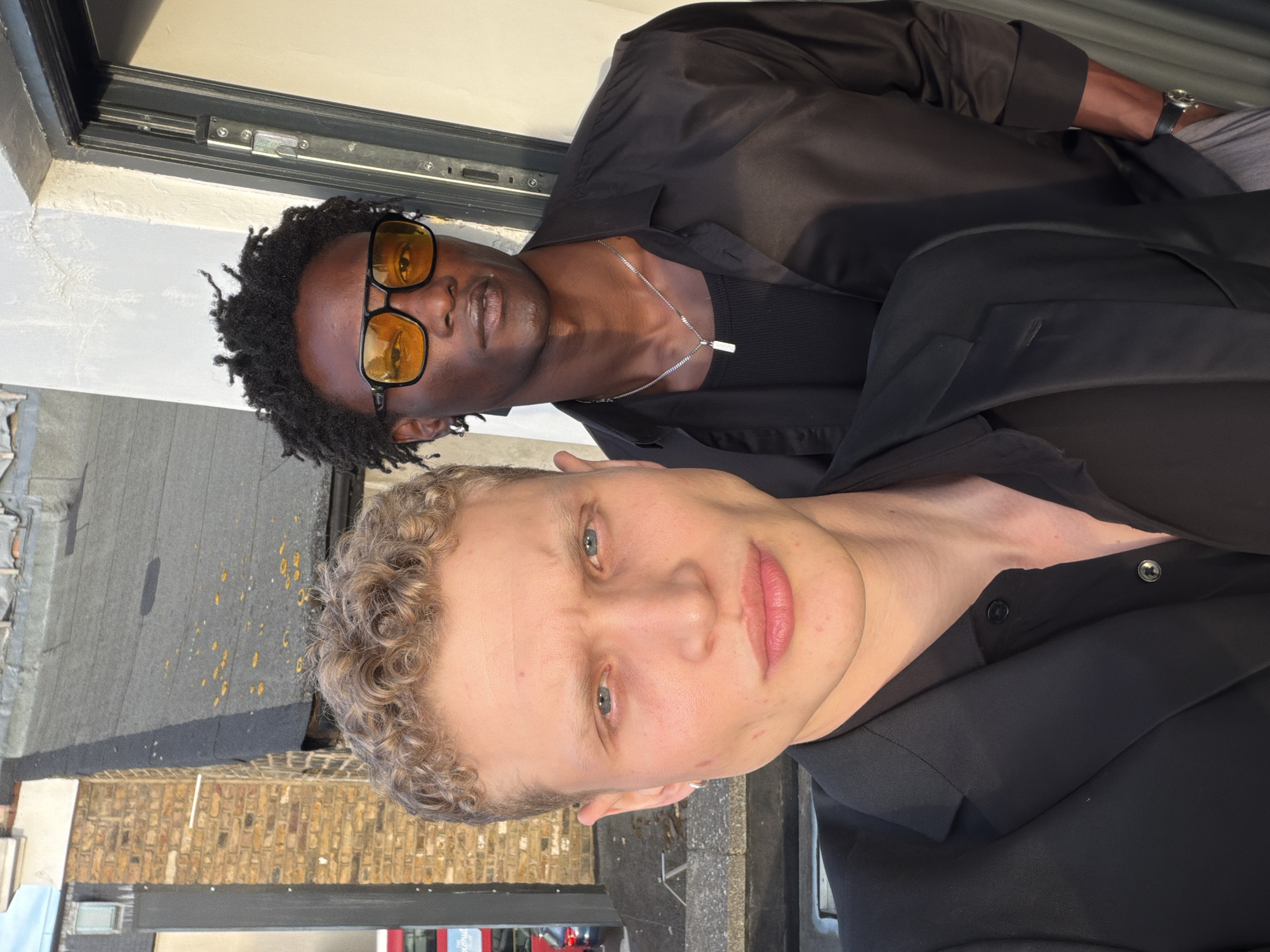 Two men are depicted in an outdoor urban setting, likely a rooftop, in the city of London, United Kingdom.
The man in the foreground, positioned slightly to the left, has light skin, blue eyes, and short, curly blonde hair. He wears a silver hoop earring in his left ear. His expression is neutral as he looks directly into the camera. He is dressed in a black blazer over a black collared shirt, which is unbuttoned at the top.
Behind and to the right of the first man stands a second man with dark skin and short, dark, textured hair. He wears square-framed sunglasses with bright yellow-orange tinted lenses, obscuring his eyes. He also maintains a neutral expression and looks directly at the camera. His attire consists of a black, long-sleeved button-up shirt worn open over a black ribbed tank top. A silver chain necklace with a small rectangular silver pendant hangs around his neck. A dark watch with a metallic face is visible on his left wrist, and his right hand is casually placed in his pocket.
The background on the left features a light brown brick building with a dark, flat rooftop exhibiting scattered yellow-orange biological growth. A partial view of a red double-decker bus is visible at street level below the building, confirming the London location. On the right, a textured white concrete wall features a dark metal door frame and an open door jamb. The scene is illuminated by strong natural light, casting subtle shadows and highlighting the subjects.