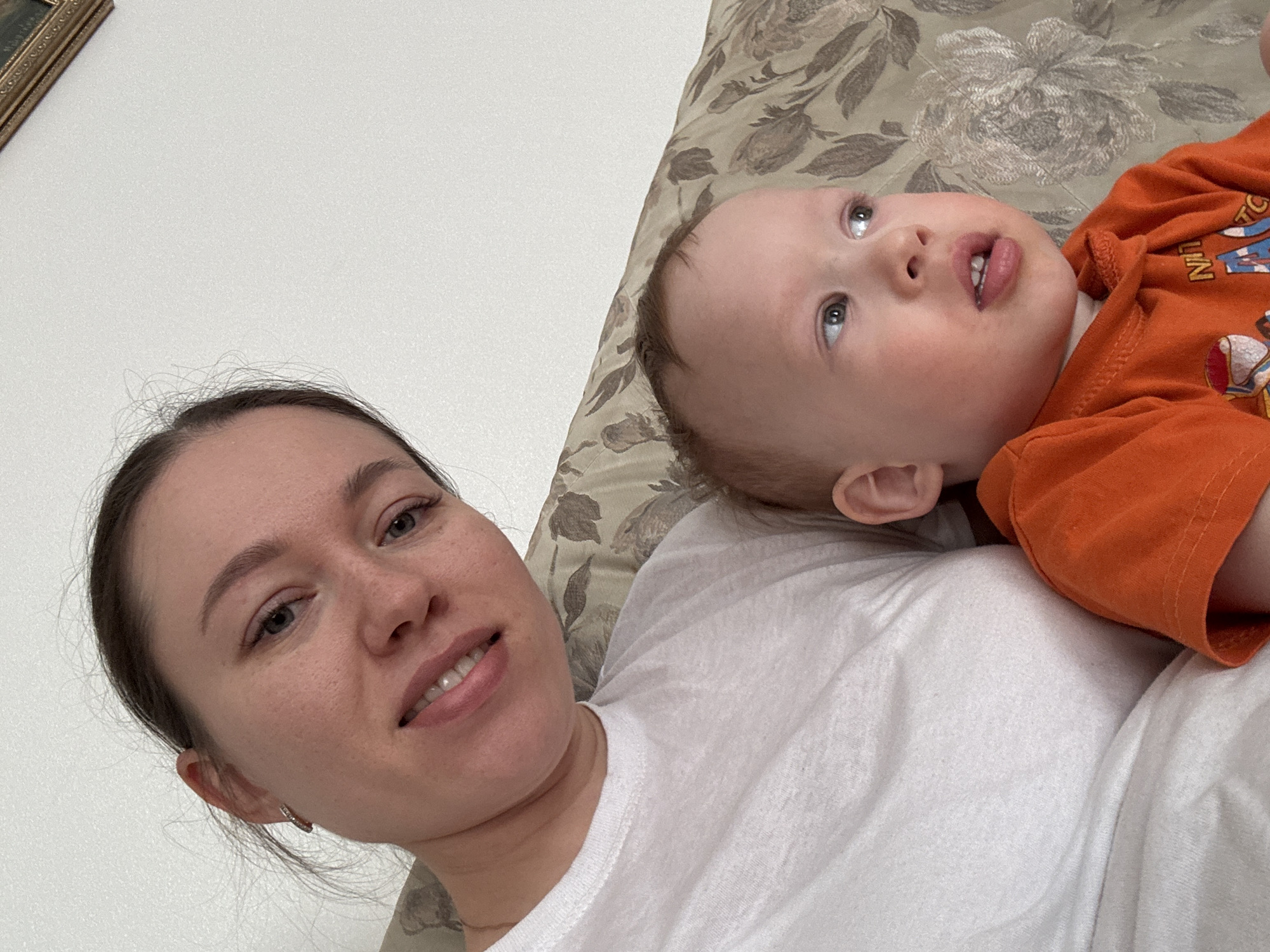 A woman and a baby are lying on a bed or couch. The woman, on the left, has dark hair pulled back from her face, and is wearing a white t-shirt. She is looking at the camera and smiling. The baby, wearing an orange shirt, is lying on the right side of the frame and looking upwards. The background consists of a floral-patterned pillow or headboard and a plain white wall on the left side. Location is unknown. The city name mentioned is irrelevant because there's no data to place them there.