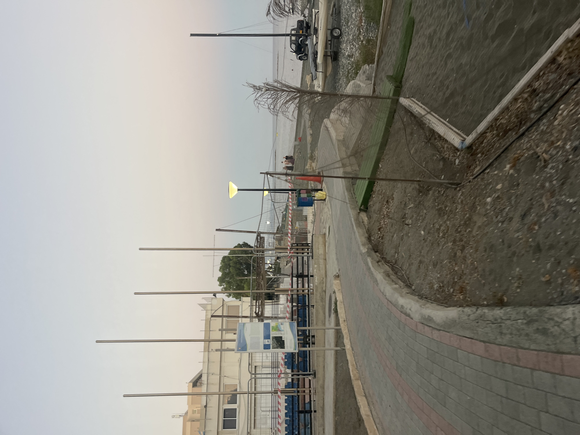 The image depicts a seaside area in Agios Tychona Community, Cyprus, at dusk. A paved walkway curves through the scene, bordered by a concrete curb and a sandy area. In the background, a building with a light-colored facade and multiple windows is visible, along with bleachers and fencing. Several tall, slender poles stand in a row, some of them likely flagpoles. The sea stretches out in the distance under a pale, fading sky. On the beach, a small boat on a trailer and a black pickup truck are parked. A street lamp with a lit yellow globe illuminates part of the pathway. There are a few people visible on the beach in the distance. The overall impression is of a public recreational area by the coast.