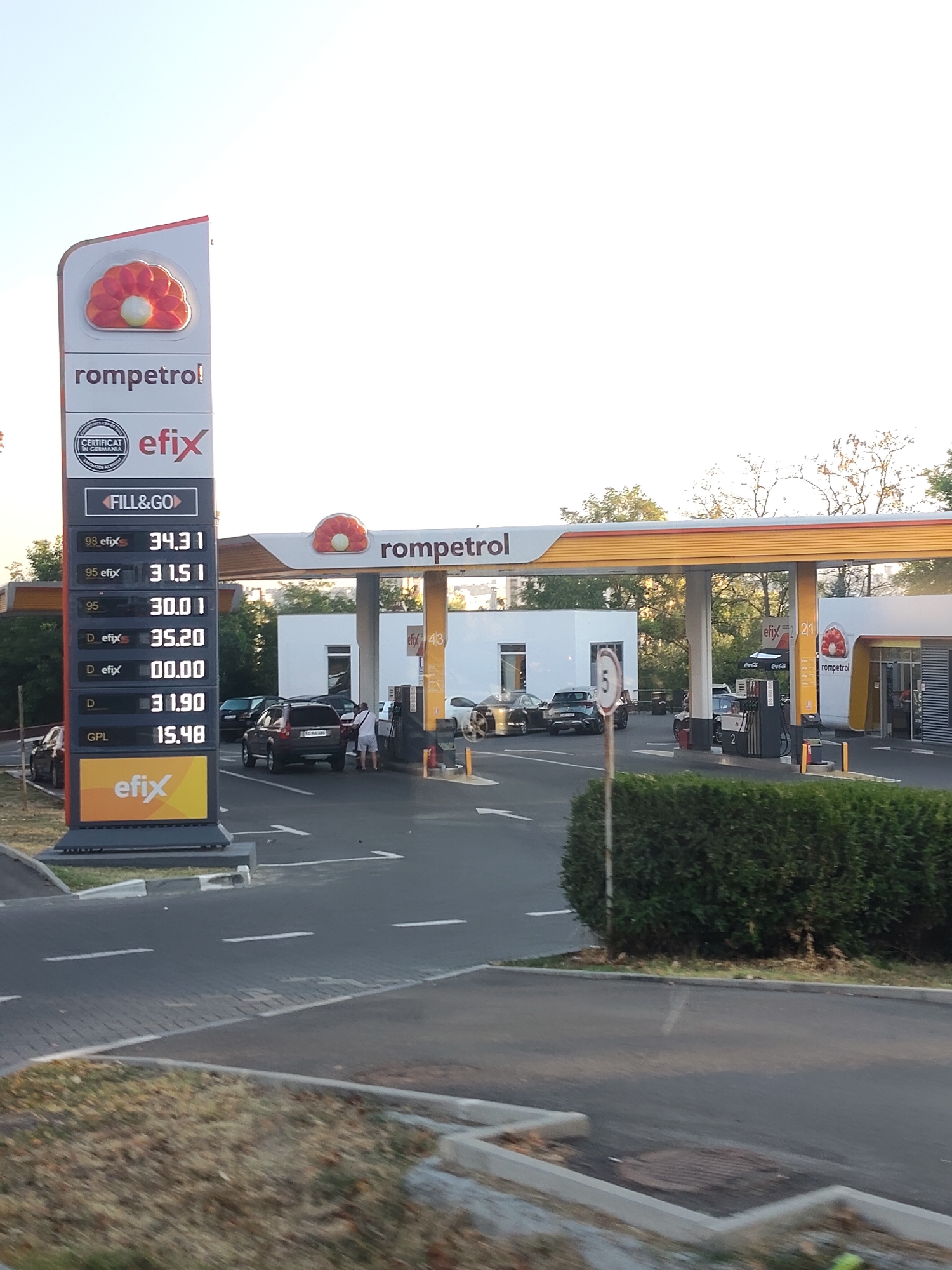 The image shows a Rompetrol gas station in Chișinău, Moldova. A tall sign displays fuel prices, with the highest price being 35.20 for D efix. Several cars are parked at the pumps. One car is being refueled by a man in a white shirt. Another person is standing near the pumps, also appearing to be a customer or employee. The gas station has a covered area with yellow accents and the Rompetrol logo. A white building with glass windows is visible behind the covered area. The scene is outdoors during daylight, with trees and bushes in the background. The ground is paved, and there are white lane markings on the road.