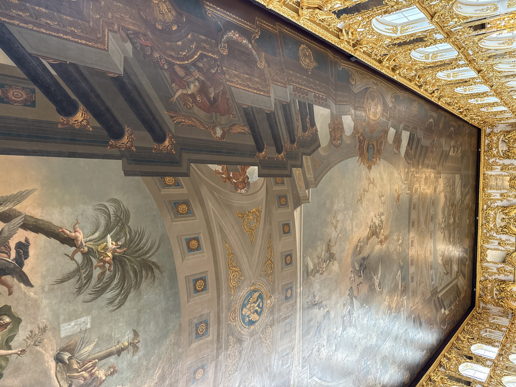 This image depicts the ornate ceiling and upper walls of a grand interior space, likely a palace hall, in Pushkin, Russia, specifically the Catherine Palace. The ceiling is a vast, frescoed expanse filled with mythological scenes and figures, painted in soft blues, grays, and ochres, with dramatic chiaroscuro. Cherubs and winged figures are prominently featured, some appearing to be suspended from or interacting with palm trees. Architectural elements like faux columns and cornices are painted to create an illusion of depth.
Below the frescoed ceiling, the upper portion of the walls is visible, adorned with richly gilded ornamentation. This gilded décor features intricate scrollwork, floral motifs, and carved figures, creating a lavish and opulent effect. Arched windows with white frames are interspersed within this gilded structure, allowing natural light to enter the space. The overall impression is one of immense grandeur and artistic mastery, characteristic of Baroque and Rococo palace interiors.