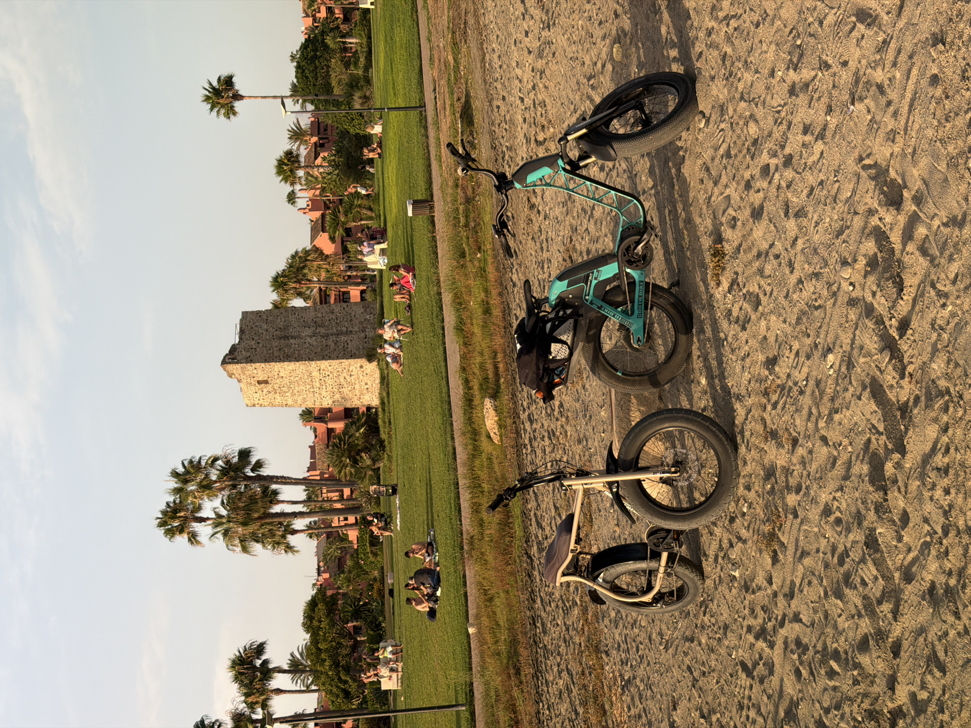 The image depicts a sunny outdoor scene on a sandy beach, with a grassy park area in the background. Two electric bicycles, one beige and one teal, are parked on the sand in the foreground. The beige bicycle on the left has a wide seat and thick tires. The teal bicycle on the right is a scooter-style electric bike with a frame that resembles a stylized triangle, also with thick tires.
In the background, on the grassy park, several people are seen relaxing and picnicking on blankets. A stone tower, possibly a historic landmark, stands prominently in the middle ground, surrounded by palm trees and reddish-orange buildings. The sky is clear with a few wispy clouds, indicating it is likely late afternoon or early evening due to the warm lighting. The overall setting suggests a leisurely day in El Velerín, Spain.