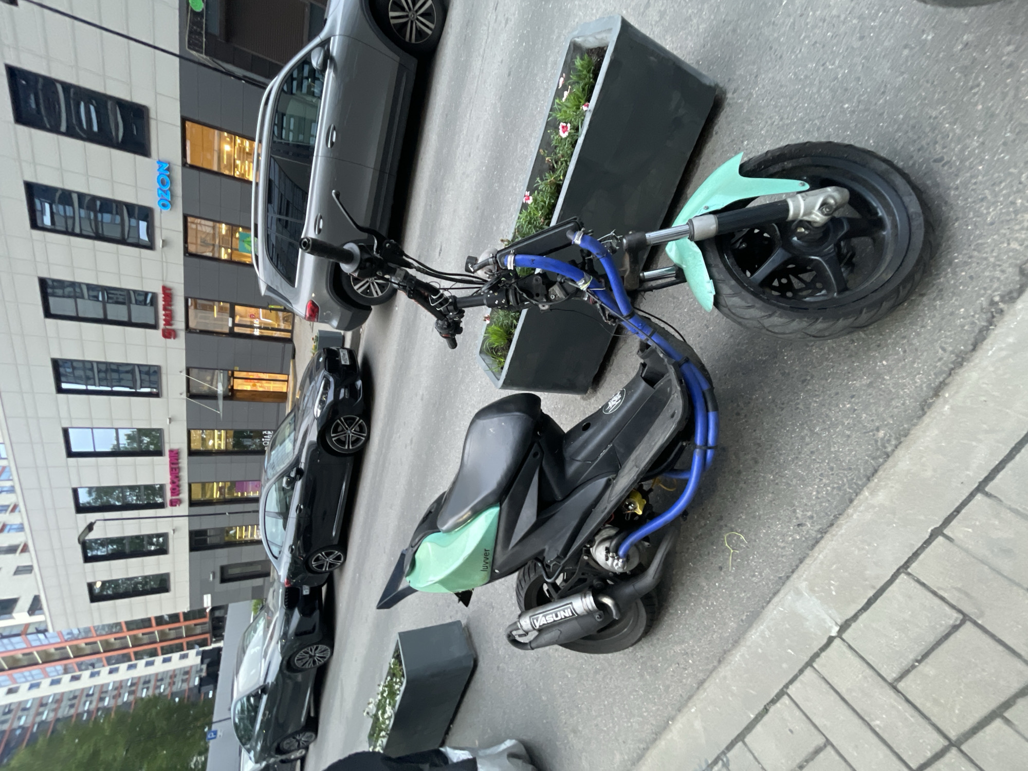An eye-level, medium outdoor shot shows a teal and black scooter parked on an asphalt street in front of a modern building with large windows. The scooter is positioned diagonally from the lower right to the upper left of the frame. The building facade is beige with dark framed windows. Signs on the building include "МАГНИТ" in red and "OZON" in blue. Several dark-colored cars are parked on the street behind the scooter. In the foreground, to the left, is a concrete planter box with white flowers and greenery. To the right, a second concrete planter box contains small pink and white flowers and green leaves. The scooter has a black seat, black handlebars with blue wires, a teal front fender, and a prominent silver exhaust pipe labeled "YASUNI." The street is paved with asphalt, and a sidewalk made of grey rectangular pavers is visible to the left. The overall lighting suggests it is daytime.