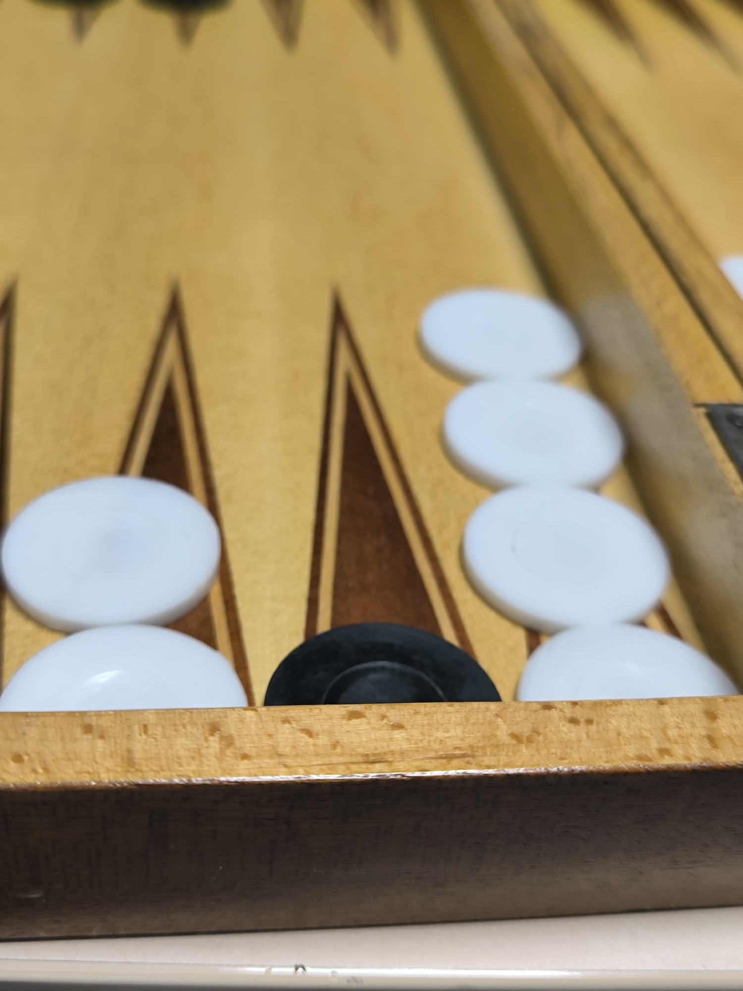 A close-up shows a backgammon board with white and one black checker pieces stacked on a point. The board is wooden with brown triangular markings on a light brown surface. The image focuses on the lower part of the board, with the checkers partially obscured by the frame. The setting is indoors. The location is unknown, and there is no evidence to suggest it is Sofia, Bulgaria.