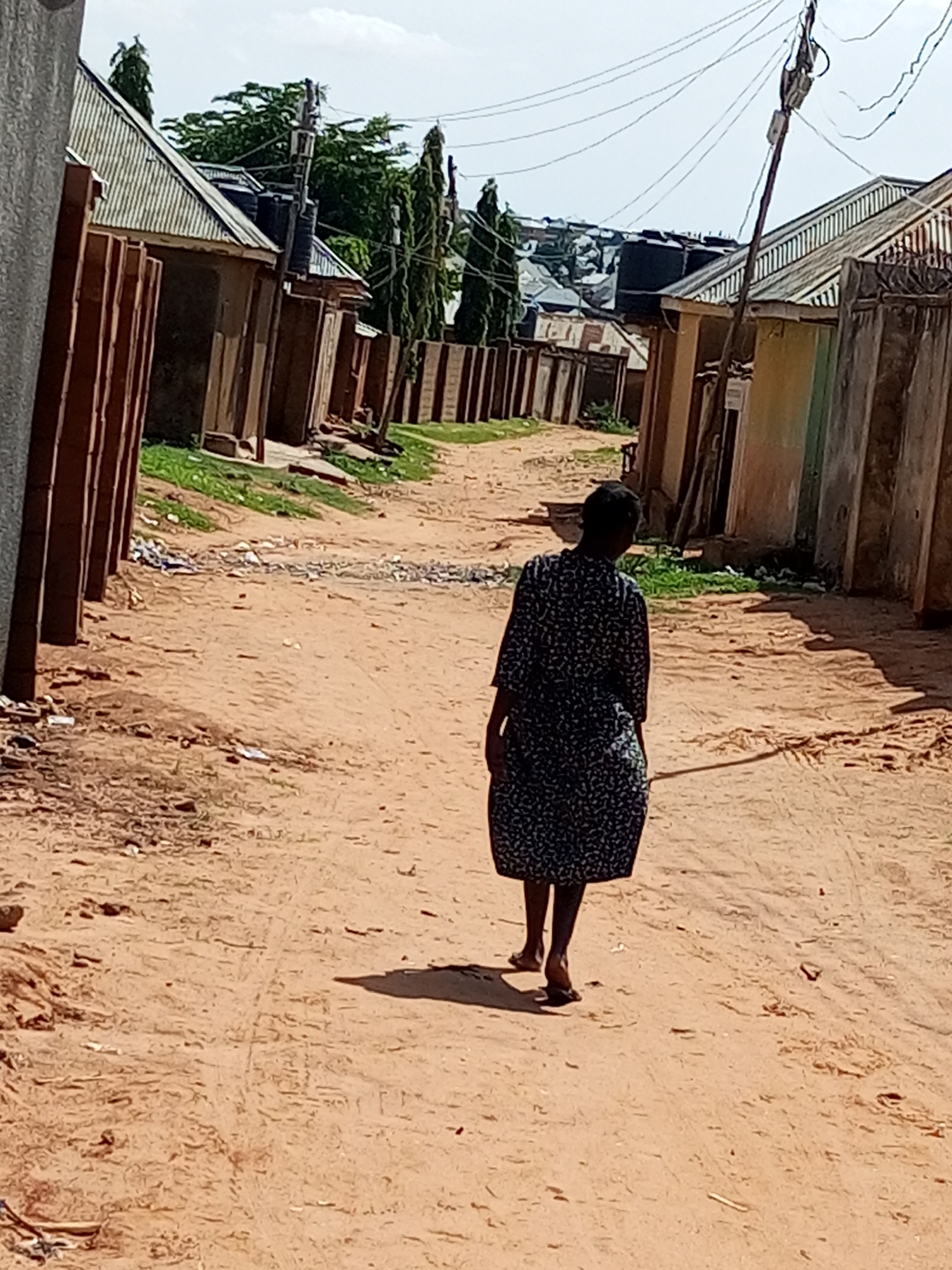 A person walks down a sandy, unpaved road between two rows of buildings. The person, who appears to be a woman wearing a dark, patterned dress, walks away from the camera. The buildings on both sides of the road are low-rise structures with corrugated metal roofs and some have fences. There are electrical wires strung across the sky above the road, supported by poles. The ground is a reddish-brown sand with some scattered debris. Green vegetation is visible in patches alongside the buildings. The sky is cloudy.