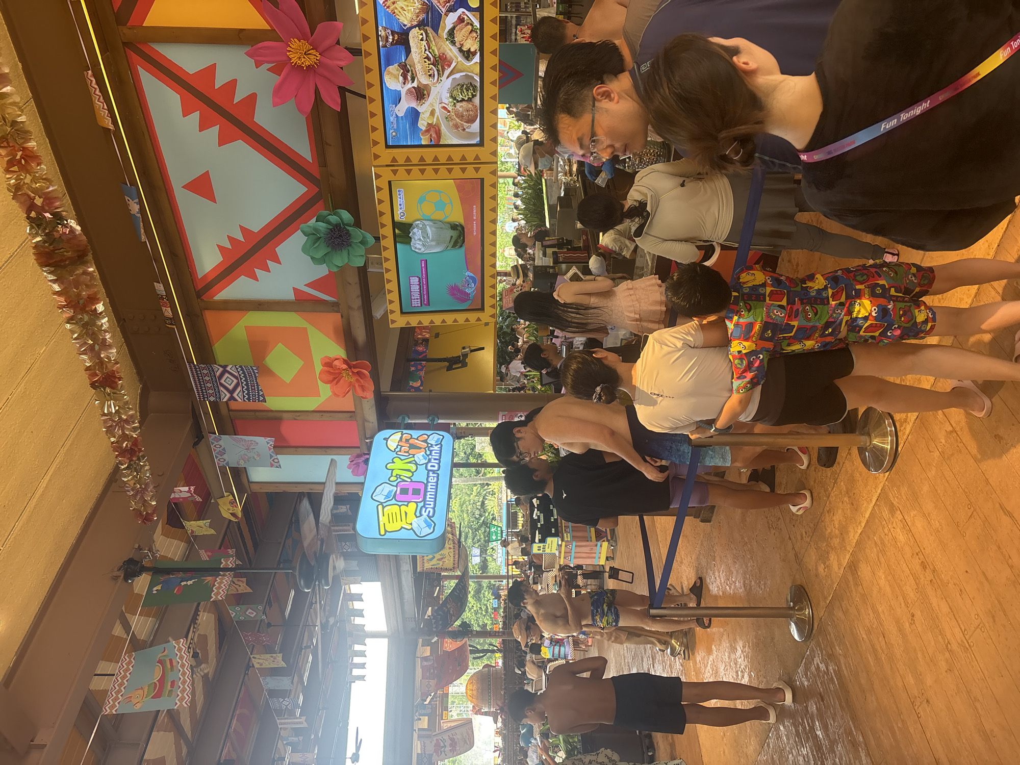 A busy outdoor food and drink service area in Panyu District, China, features a long queue of people waiting to order. The area is decorated with colorful banners and large paper flowers, giving it a festive, tropical theme. Several overhead fans are visible, suggesting a warm climate.
In the foreground, a blue rope barrier divides the waiting line. People in the queue are dressed in casual summer attire, many in swimsuits, indicating a water park or beach resort setting. A young boy in the line wears a vibrantly colored romper featuring cartoon characters.
Above the queue, a bright blue sign with Chinese characters and the English text "Summer Drink" is prominently displayed, along with an image of iced drinks. To the right, two large digital screens showcase menu items. The left screen displays a drink with ice and a soccer ball graphic, labeled "Pandam Extract with Coconut Milk." The right screen shows an assortment of food, including burgers, sandwiches, fries, and other dishes.
The background reveals more of the establishment, with patrons seated at tables and lush greenery visible through open structures, suggesting a large entertainment complex. The wooden flooring and the overall ambiance point towards a recreational facility designed for summer enjoyment.