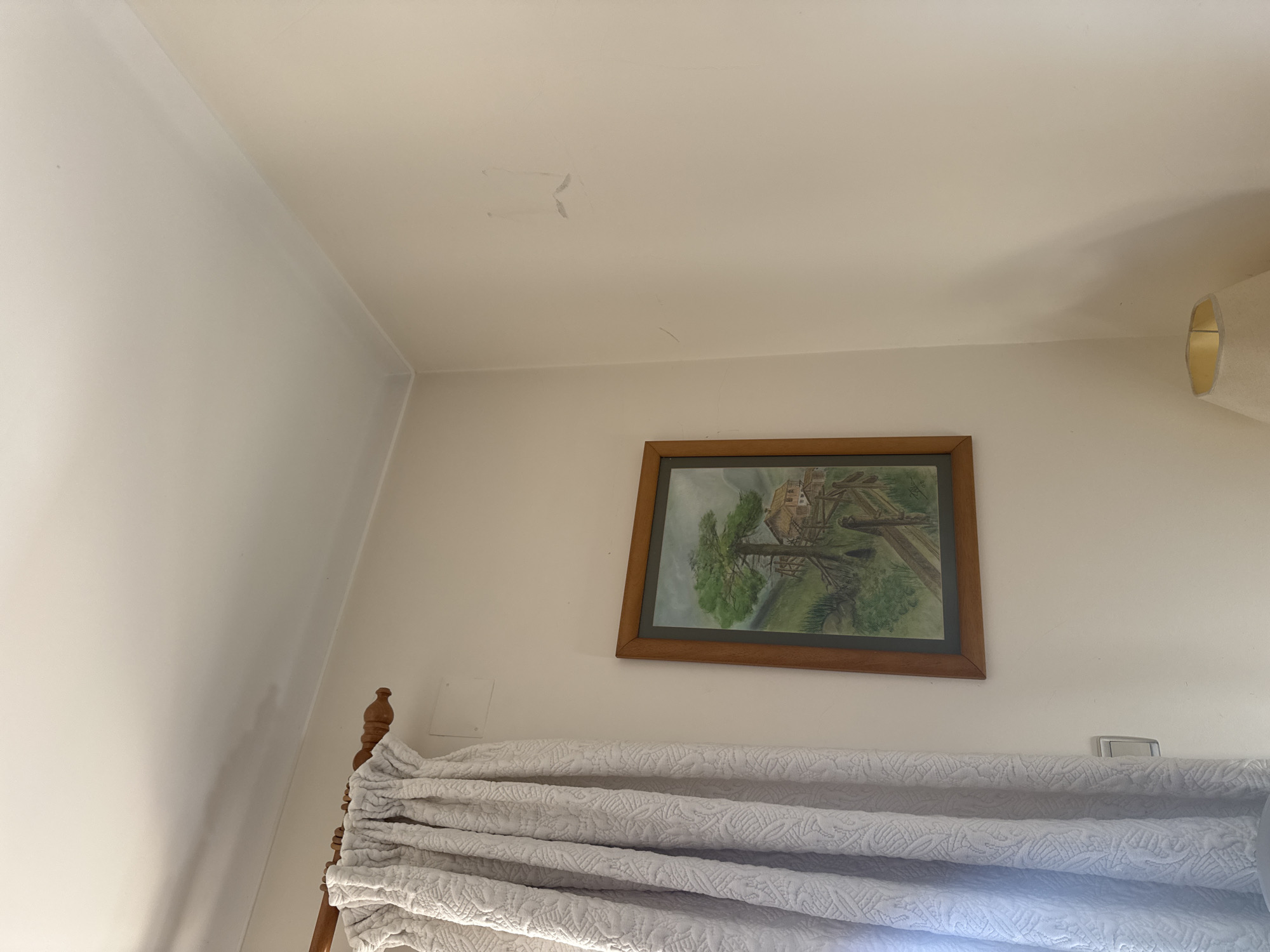 The scene depicts an interior corner of a room located in Mirandela, Portugal. The walls are painted a light beige or off-white color, meeting a white ceiling with a simple cornice molding.
On the left side of the frame, a textured light-colored curtain, possibly off-white or light grey with a raised patterned design, hangs from a wooden rod. The wooden rod is light brown and features a rounded finial on its visible left end. A square white electrical access panel is visible on the wall above the midpoint of the curtain. Partially visible near the bottom left, behind the curtain, is a light switch.
Centered on the left wall, a rectangular framed artwork is displayed. The frame is dark brown wood. The artwork itself is a landscape painting featuring a prominent green-leafed tree, a rustic wooden house or structure, and a wooden fence situated on a green hillside under a light sky.
The right wall is largely bare, exhibiting minor scuff marks and faint, light-colored streaks, including a distinct U-shaped mark towards the upper right quadrant. In the bottom right corner, the upper portion of a table lamp with a light beige fabric lampshade is visible. No individuals are present in the scene.