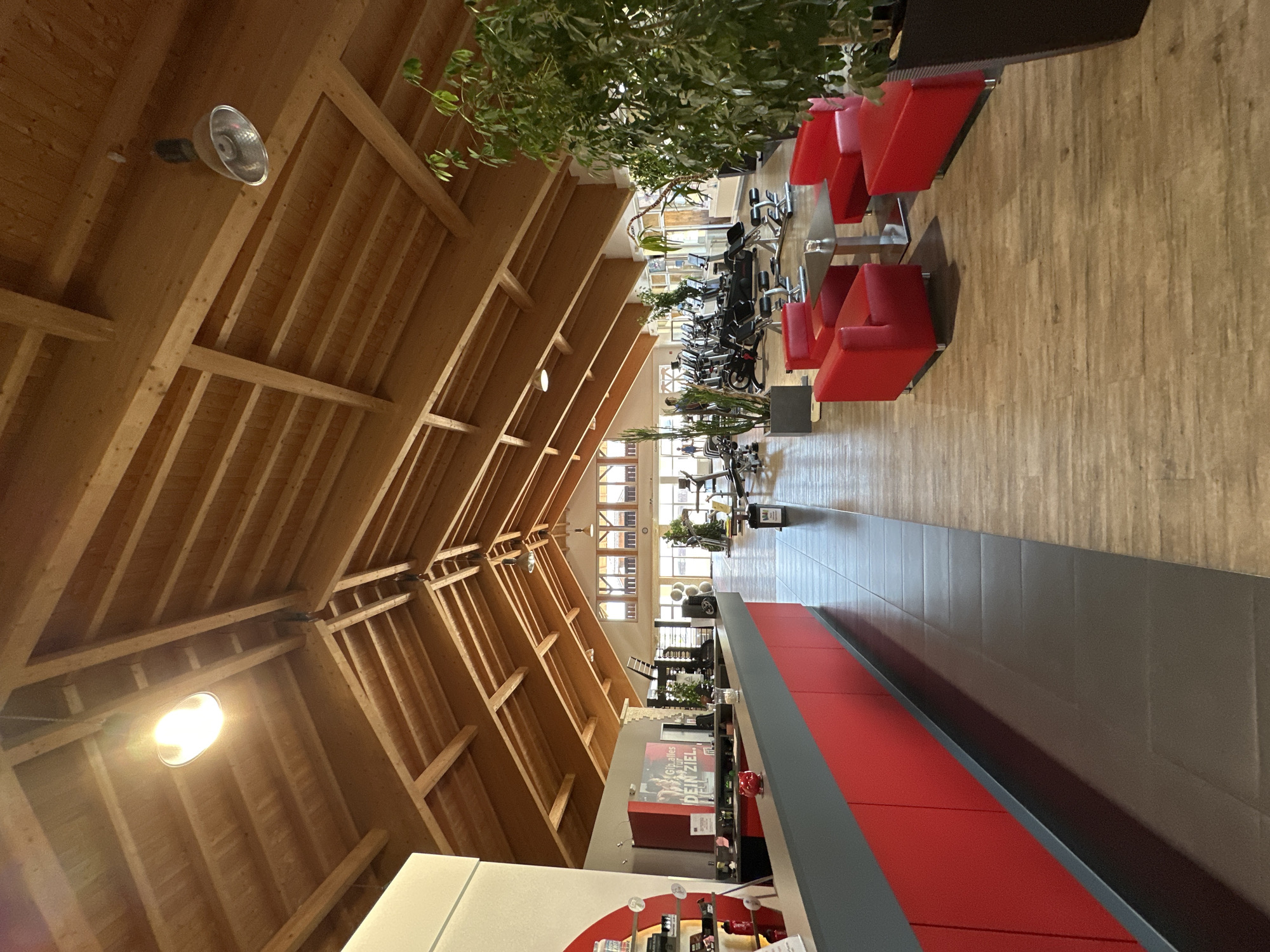 The image shows the interior of a fitness studio. A reception desk with red cabinets and a grey countertop is in the foreground on the left. Behind the desk, a sign with German text "Glaub. Alles für DEIN ZIEL." is visible. The studio has a high, pitched wooden ceiling with exposed beams and several lights. The floor is a mix of dark tiles near the reception and light wooden planks further in.
In the mid-ground and background, various exercise machines, including treadmills and elliptical trainers, are arranged in rows. Two people are visible in the distance near the exercise equipment. To the right of the exercise area, there is a seating area with red armchairs and a black coffee table. A large potted plant with lush green foliage is situated in the far right of the image. Natural light streams in from windows at the far end of the studio, which appear to overlook an outdoor area with more wooden structures, possibly part of a building. The overall impression is that of a modern, well-lit fitness facility. The provided information states the location is Garmisch-Partenkirchen, Germany.