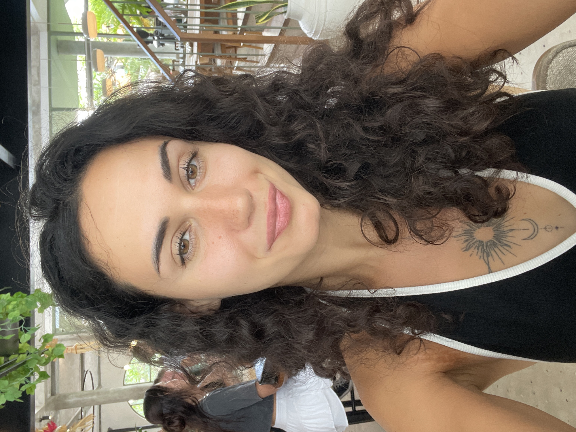 A medium shot shows a woman with dark curly hair and light skin with freckles taking a selfie in an indoor restaurant. She is looking at the camera and smiling with her lips slightly parted. She is wearing a black tank top with white trim and a tattoo on her chest. In the background, there are other people, tables, chairs, and plants. The overall lighting is natural and bright, suggesting it is daytime. The location is identified as a restaurant in North Kuta, Indonesia.