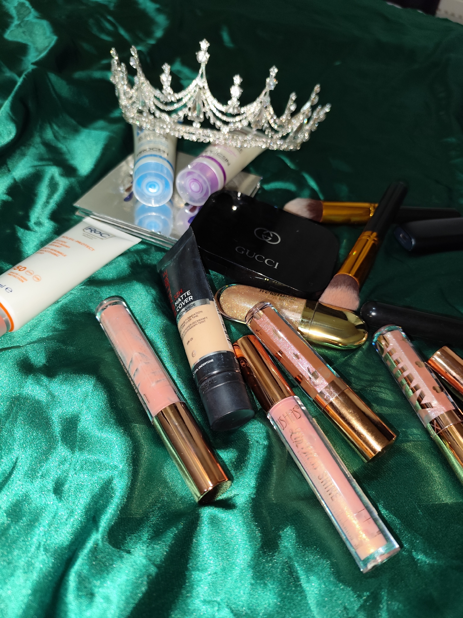 The image shows a variety of beauty products arranged on a green, shimmering fabric. Dominating the scene is a silver crown.
Visible cosmetic items include:
* Tubes of ROC sunscreen
* L'Oréal Matte Cover foundation
* Assorted lip glosses
* Gucci compact
* Makeup brushes
Additional details are the reflections of some of the items, like the tubes, on a reflective surface, which appears to be a mirror.
There is no information in the image to indicate that this scene is taking place in Sarıyer, Turkey.