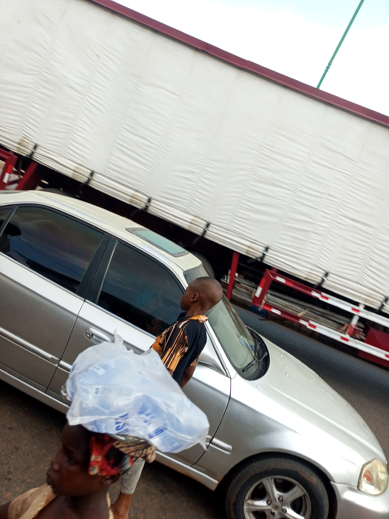 In an outdoor urban setting, a silver sedan is parked next to a large truck with a white tarp-covered trailer. The truck has red and white reflective markings on its sides. A young boy wearing a black and orange patterned shirt stands between the car and the truck, looking towards the truck. In the foreground, a person with dark skin, wearing a red, black, and gold headwrap, carries a large, clear plastic bag filled with what appears to be water bottles on their head. The image is taken from a slightly elevated angle, with the sky visible at the top.