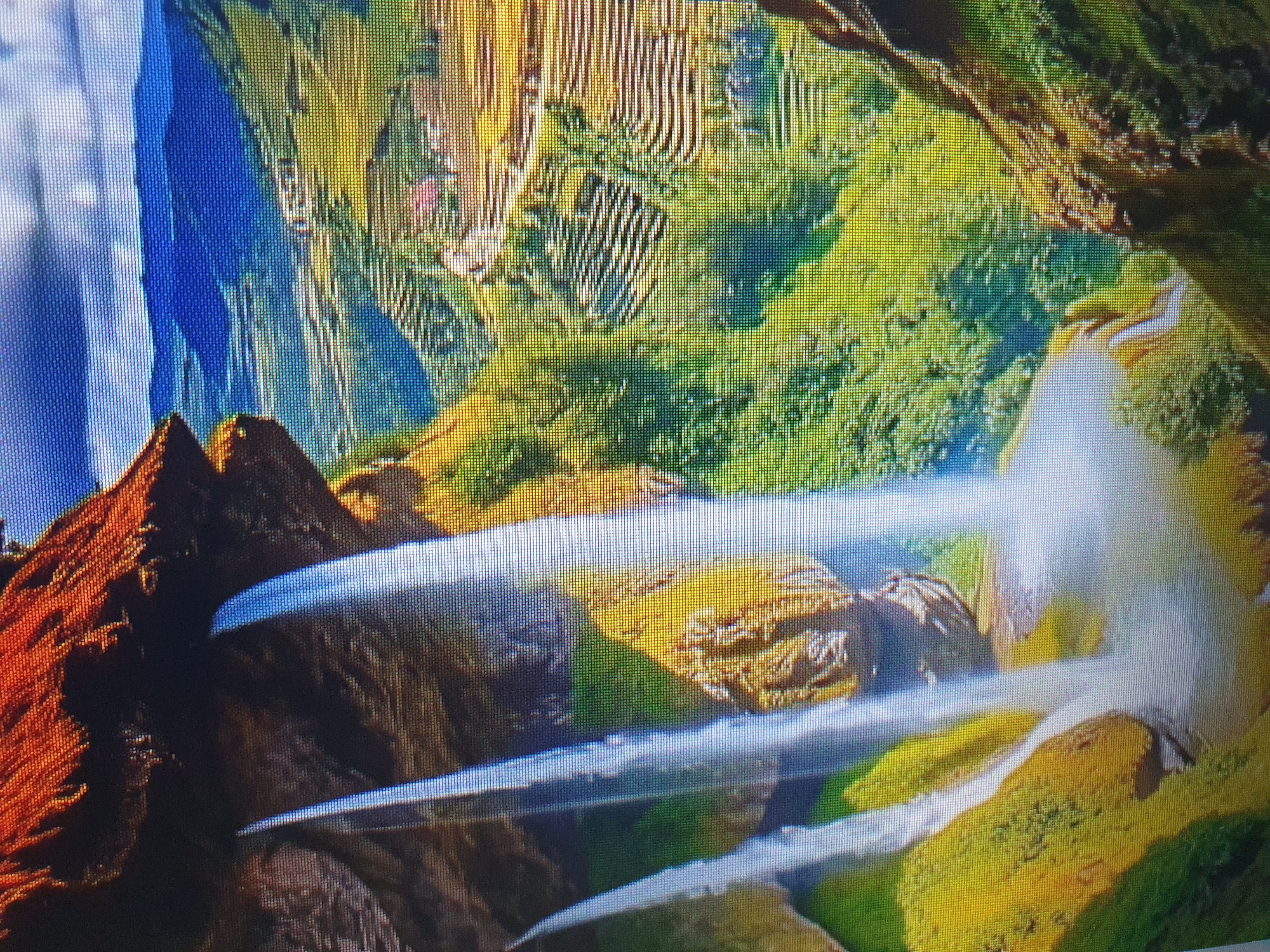 The image depicts a cascading waterfall flowing over rocky terrain covered in green vegetation and orange-brown rock formations. In the background, a valley stretches out with terraced fields, some appearing golden yellow and others green, interspersed with trees. Distant blue mountains rise under a sky with scattered clouds. This scene is located in Tashkent, Uzbekistan.