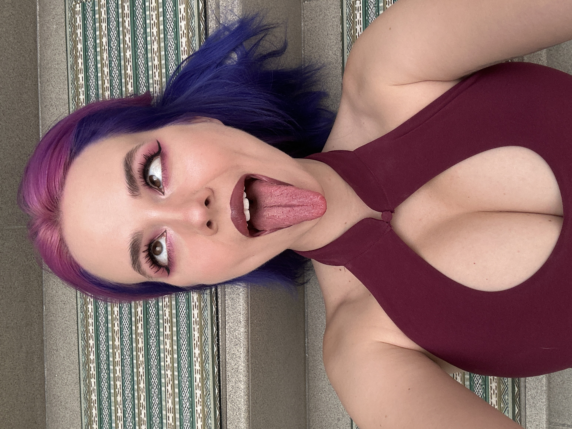 A woman with purple and blue hair is lying on a patterned surface. She is wearing a maroon halter top with a cut-out design. Her mouth is open, and her tongue is sticking out. Her makeup includes pink eyeshadow. The location appears to be indoors, in the City of Zagreb, Croatia.