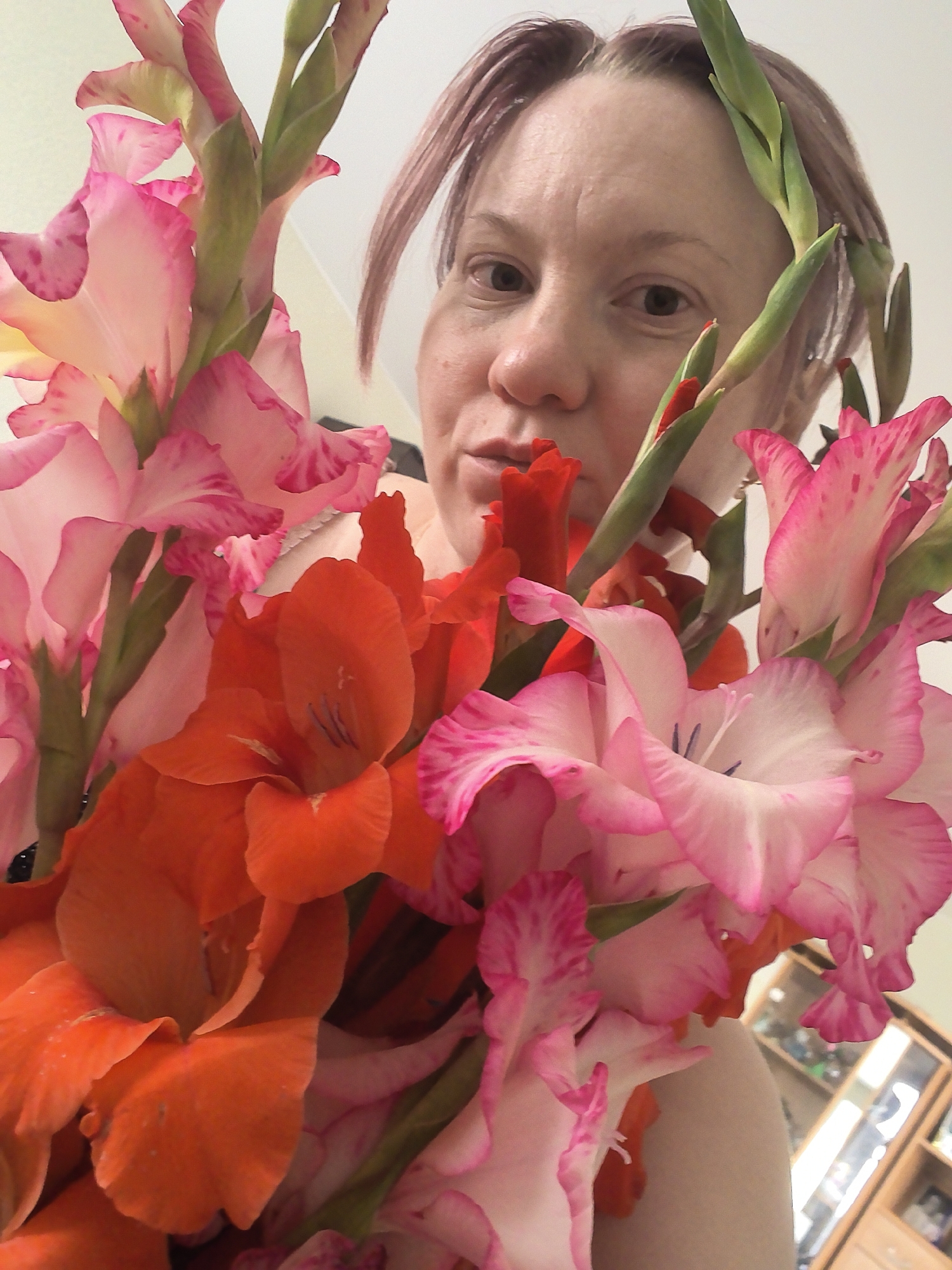 A close-up, low-angle shot features a woman holding a bouquet of gladiolus flowers in front of her face. The flowers, in shades of pink and orange, dominate the foreground. The woman, who appears to be middle-aged with light skin and short, reddish-brown hair, looks directly at the camera. Her face is partially obscured by the blossoms. Her expression is neutral. The background is out of focus, with hints of a light-colored wall and a wooden cabinet with glass doors, possibly in a domestic setting. There is no clear indication of the location within Chaikovsky, Russia.