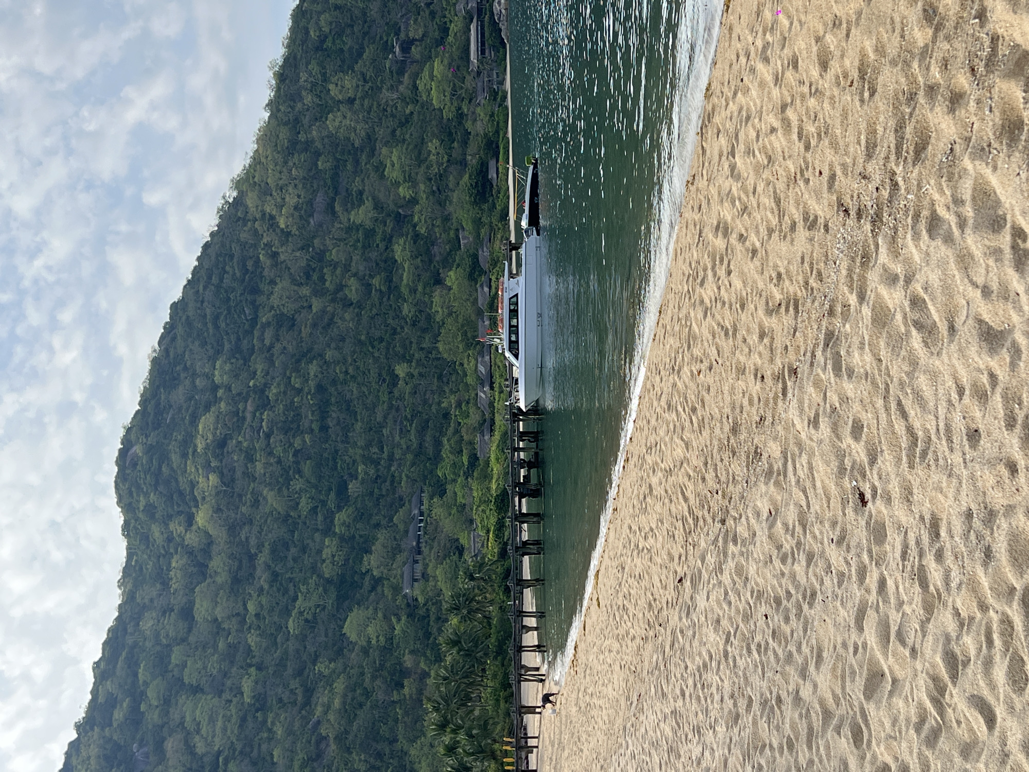 A wide, sandy beach in the foreground transitions to calm, turquoise water. A wooden pier extends from the left side of the frame into the water, with a white speed boat docked alongside it. A smaller, dark-colored boat is tethered to the speed boat. In the background, a densely forested green mountain rises, dotted with some buildings nestled amongst the trees. The sky above is a pale blue with scattered white clouds. A single black and white dog is visible on the sand near the pier. The scene suggests a tropical coastal location.