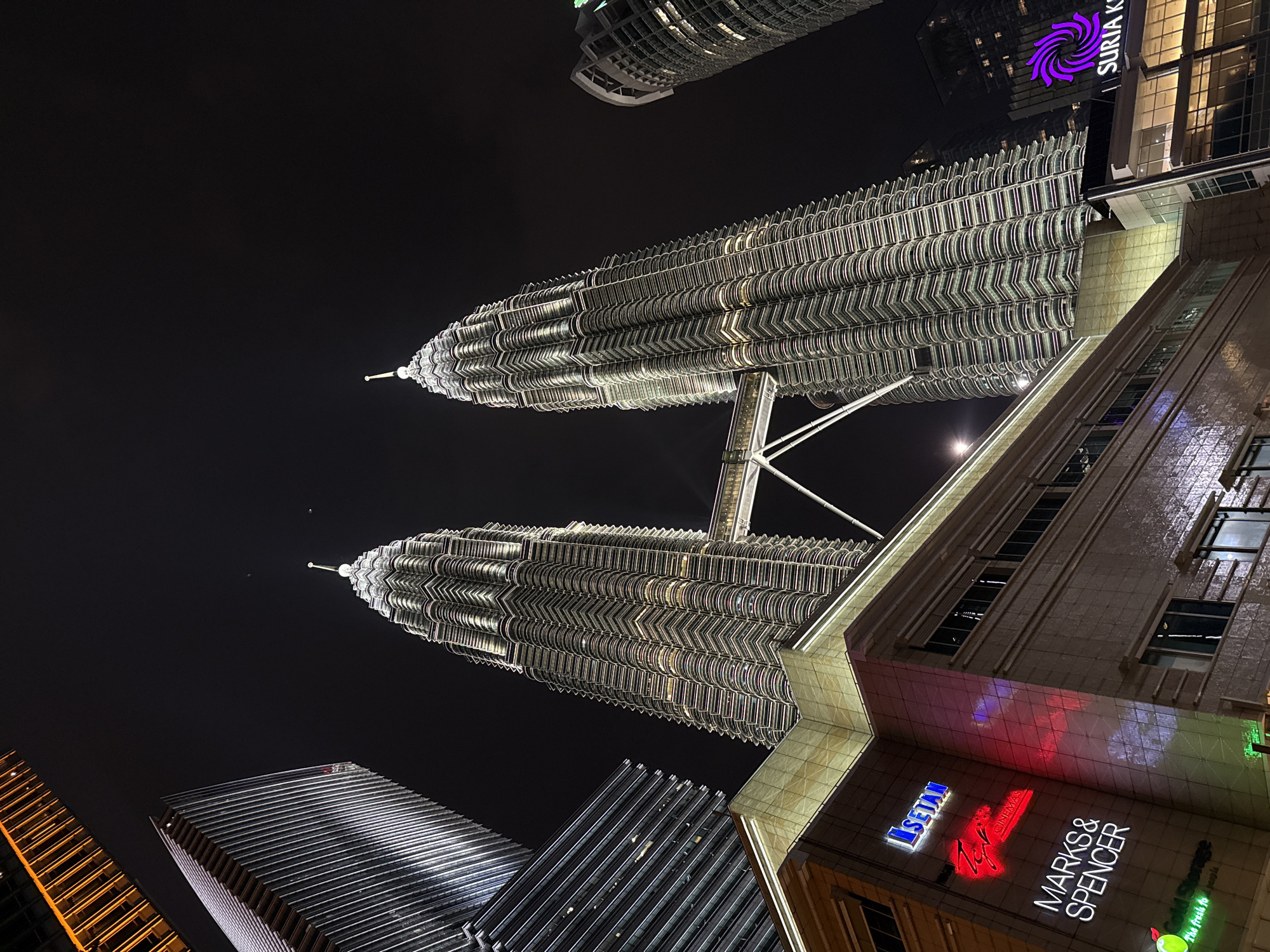The image shows the illuminated Petronas Twin Towers at night in Kuala Lumpur, Malaysia, viewed from a low angle. The towers dominate the upper portion of the image against a dark sky. In the foreground, the side of a building is visible, with the logos of "ISETAN" and "MARKS & SPENCER" illuminated. Other buildings are also present in the image.