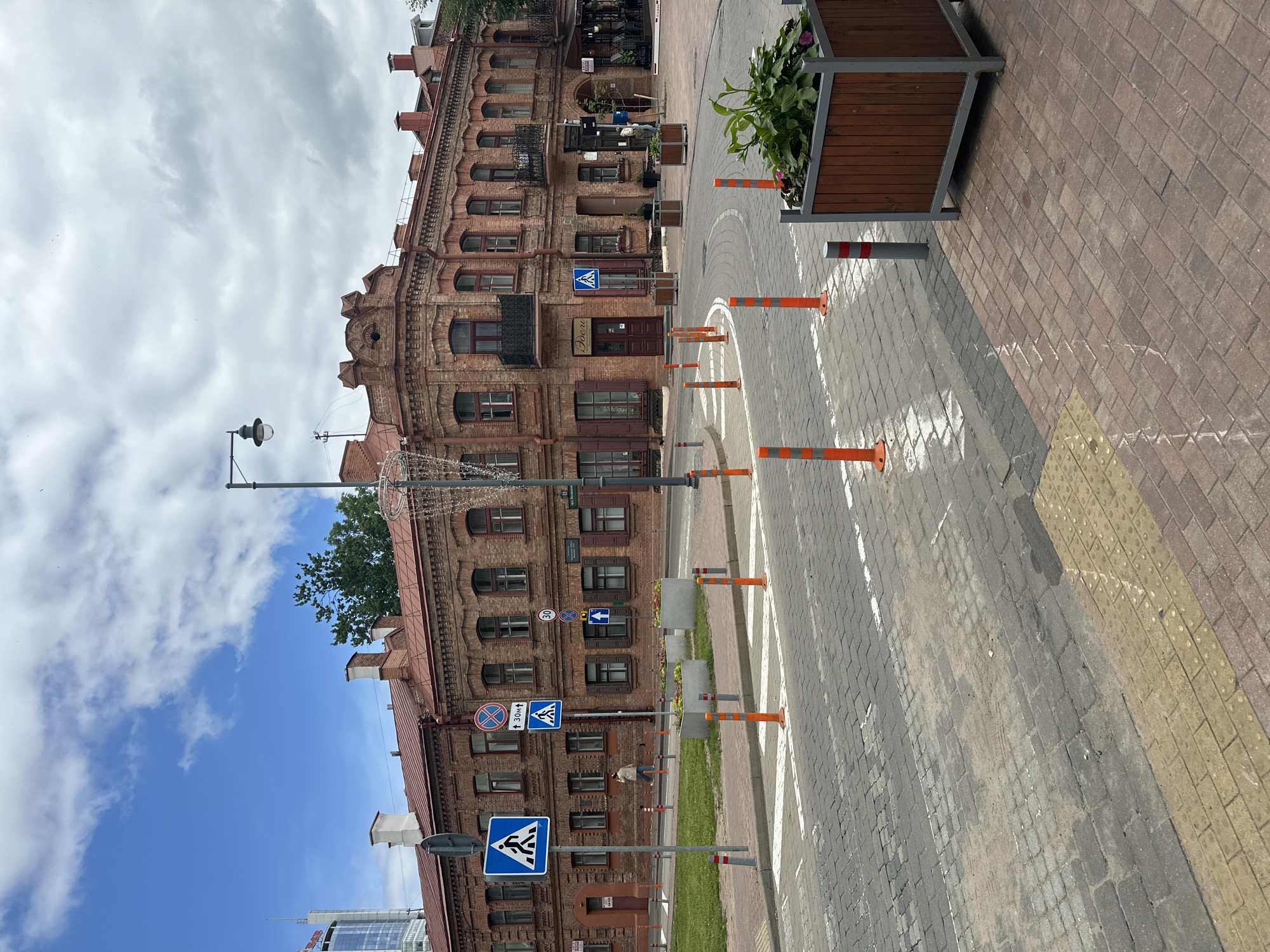 The image depicts a street scene in Minsk, Belarus, featuring a historic brick building with arched windows and decorative cornices. The building, likely from the late 19th or early 20th century, has a red tile roof and dark wooden shutters. A sign above an entrance reads "Двери" (Doors). In front of the building, a pedestrian crossing is marked with white lines on a cobblestone street, bordered by planters with greenery. Orange traffic cones and bollards are placed along the street, indicating traffic management. Several blue traffic signs are visible, including a pedestrian crossing sign and a sign with the number "30" and an arrow, possibly indicating a speed limit. A lone person is walking across the street. The sky is partially cloudy, and a modern glass skyscraper is visible in the distance on the left side of the image. The foreground shows the texture of the cobblestone pavement and a yellow tactile paving strip.