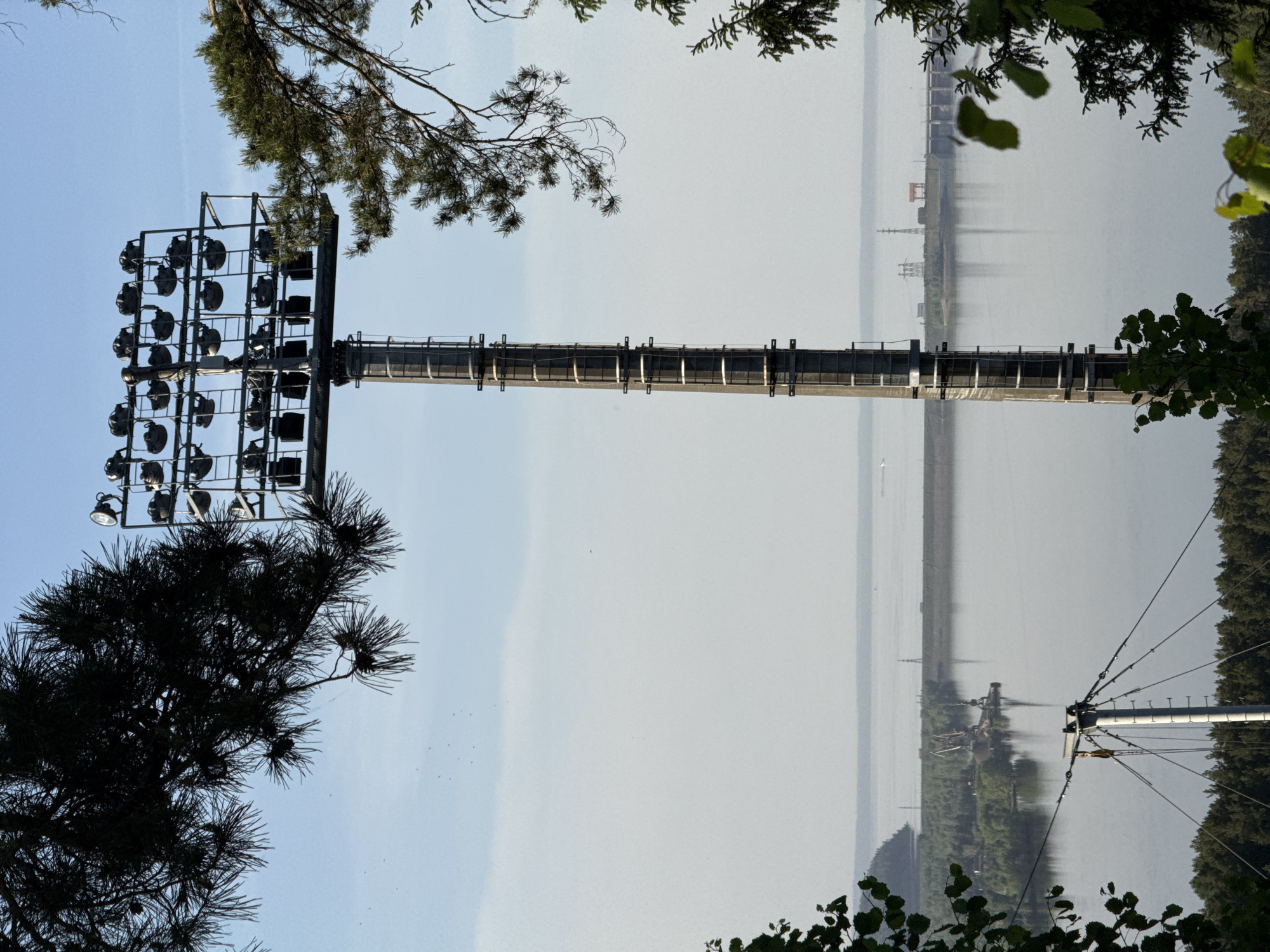 A long-range shot shows a tall lighting tower with multiple floodlights attached. The tower appears to be located near a body of water, possibly a large river or lake. The opposite shoreline is visible in the background. The surroundings are lush with vegetation, including trees and shrubbery, some of which are in the foreground, obscuring parts of the view. The image appears to be taken in the Прикамский region of Russia.