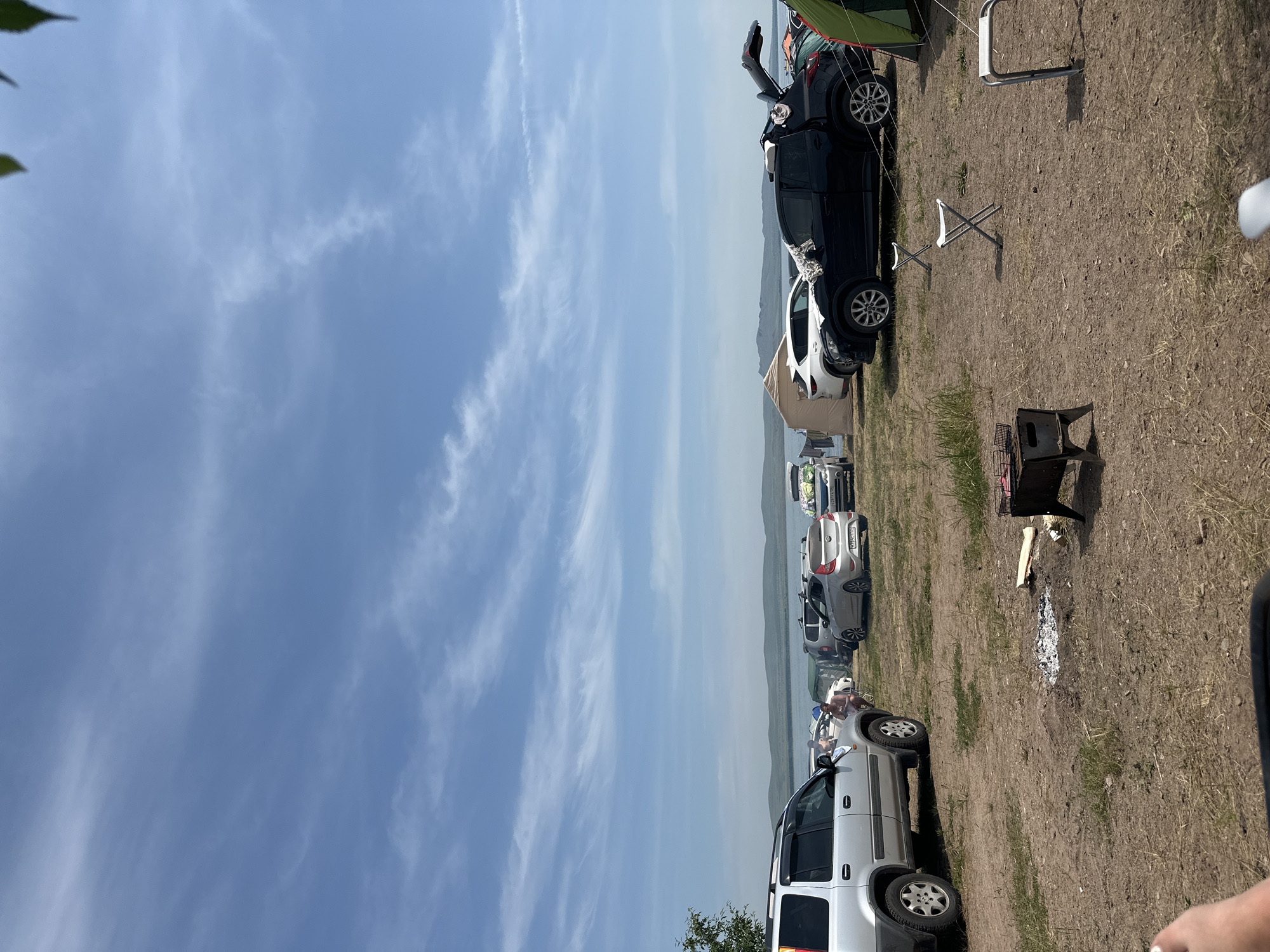 The image depicts a scenic outdoor campsite by a body of water, under a partly cloudy blue sky. The ground is dry and uneven, with patches of sparse grass. Several vehicles, including SUVs and sedans, are parked in the area, suggesting a gathering or event. A portable charcoal grill is set up with embers, indicating recent use for cooking. Three folding chairs are placed around the campsite, some of which appear empty. In the background, a tent and camping equipment are visible, alongside a distant shoreline with rolling hills. The overall atmosphere is relaxed and recreational, with a sense of people enjoying the outdoors. The location appears to be a natural landscape, possibly a park or designated camping area, with the city of Колодезный, Russia in the vicinity.