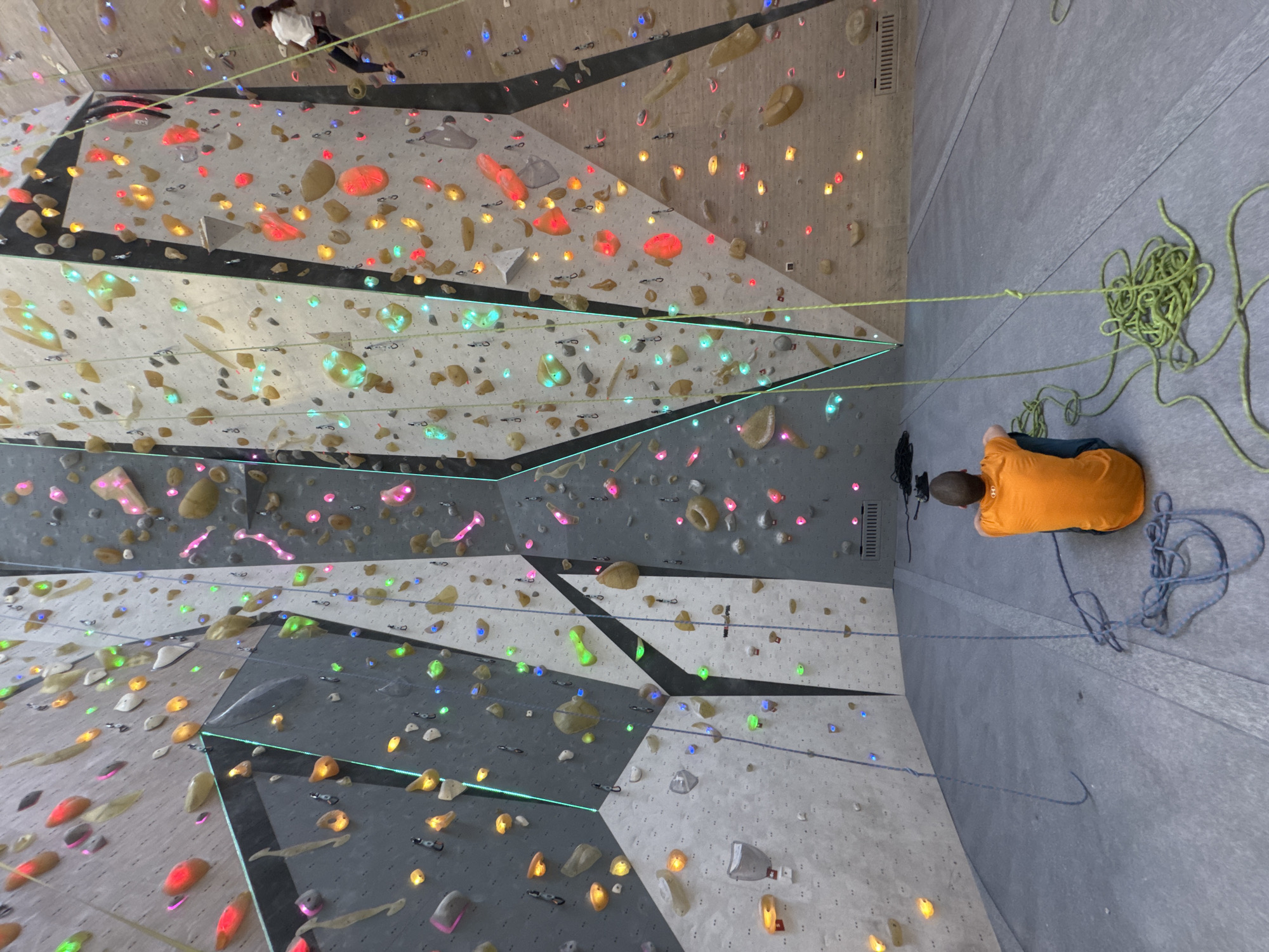 This vertical image depicts an indoor rock climbing gym, taken from a slightly elevated perspective looking up at the wall. The climbing wall is composed of large, angular panels in varying shades of light and dark grey, covered with a multitude of climbing holds. Many of these holds are internally illuminated with LEDs, glowing in a spectrum of colors including red, orange, yellow, green, blue, pink, and purple, likely indicating specific climbing routes.
In the upper left portion of the image, a person in a white t-shirt and dark pants is actively climbing the wall, appearing to ascend. On the right side of the frame, which represents the floor of the gym, another person wearing an orange t-shirt and dark bottoms is seated or kneeling. A bright green climbing rope extends from near this person up the wall, and a large coil of the same rope rests on the floor next to them, indicating they are likely belaying the climber. Another rope, possibly blue or dark grey, is visible further down the wall. The scene captures an active climbing session within a modern gym environment. There is no prominent or legible text visible in the image.