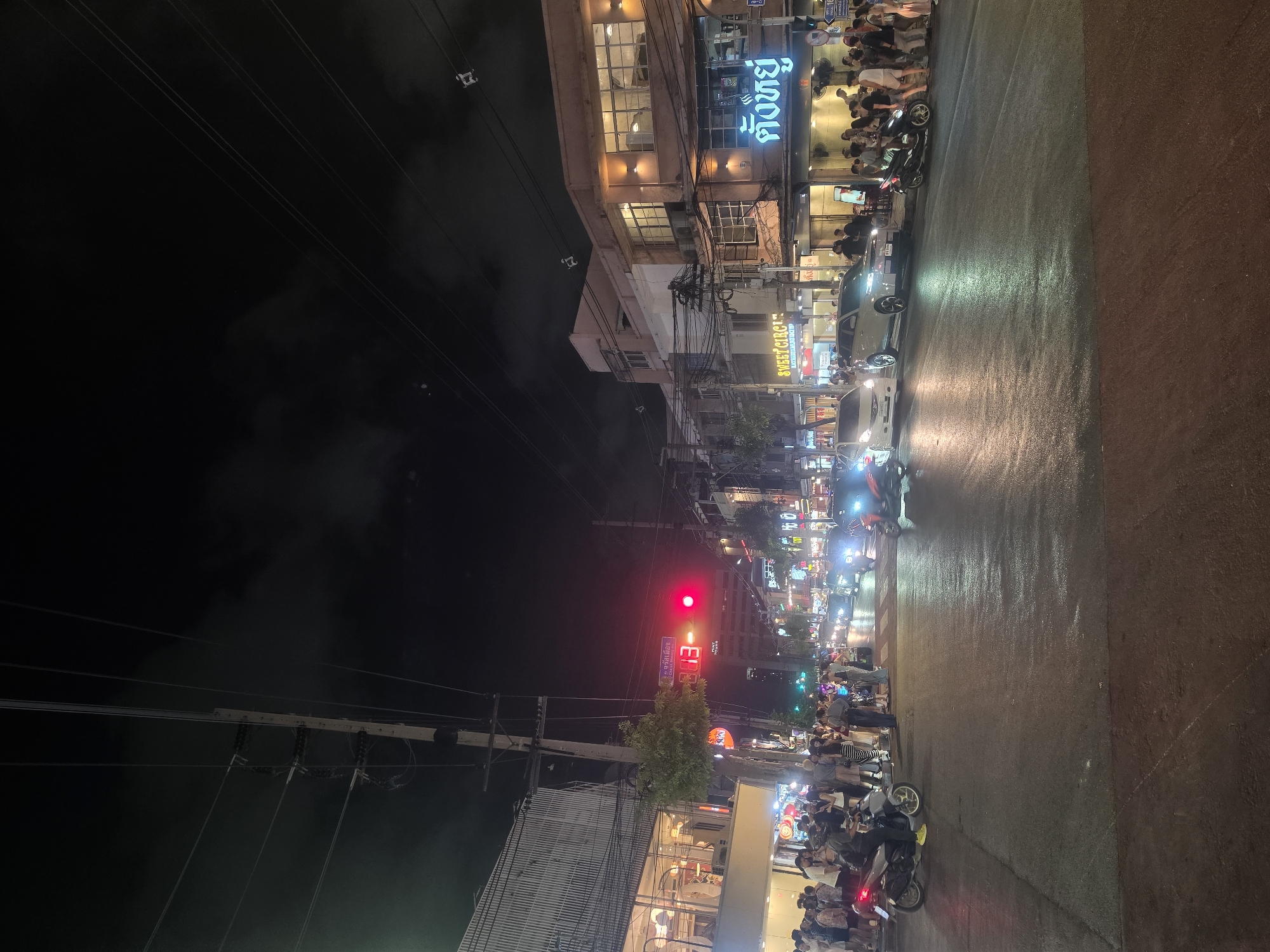 This photograph, taken at night, captures a vibrant street scene in Bangkok, Thailand. The image is rotated.
A line of pedestrians and motorcycles are waiting at a red traffic light. The road is wet, reflecting the city lights. The street is lined with buildings, some of which are illuminated with neon signs in Thai. The sky is dark. Power lines crisscross above the street. Traffic is visible in the distance.