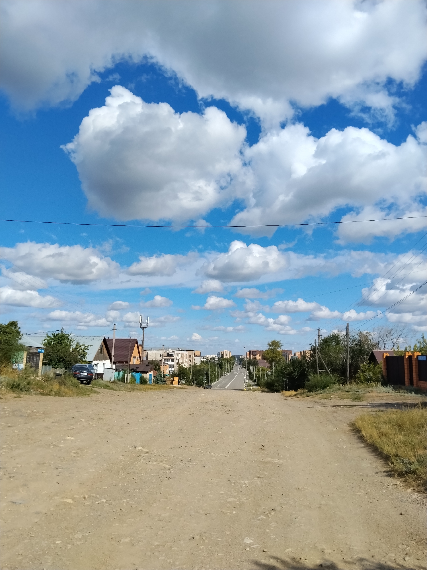The image depicts a view looking down a dirt road in a suburban area of Kokshetau, Kazakhstan. The road is unpaved and appears to be in a rural or developing part of the city. On either side of the road, there are low-rise buildings, including houses with pitched roofs and some apartment buildings in the distance. There are also trees and vegetation along the sides of the road. A dark-colored car is parked on the left side of the road. The sky is a vibrant blue with numerous fluffy white clouds, suggesting fair weather. Overhead, power lines are visible, strung between poles. The overall impression is of a quiet, residential street on a sunny day.