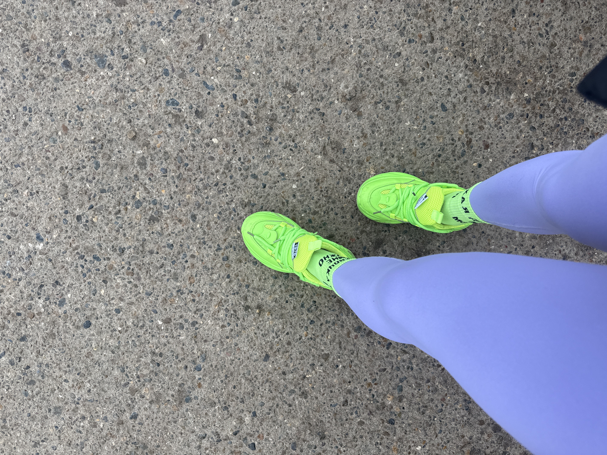 A top-down shot shows a person standing on a gray concrete surface. The person is wearing light purple leggings and neon green athletic shoes with matching neon green socks. The concrete surface is speckled with small dark-colored aggregate. No other individuals or objects are visible. The specific location within Kauchuk Stantsiya, Russia is indeterminable based solely on the provided image.